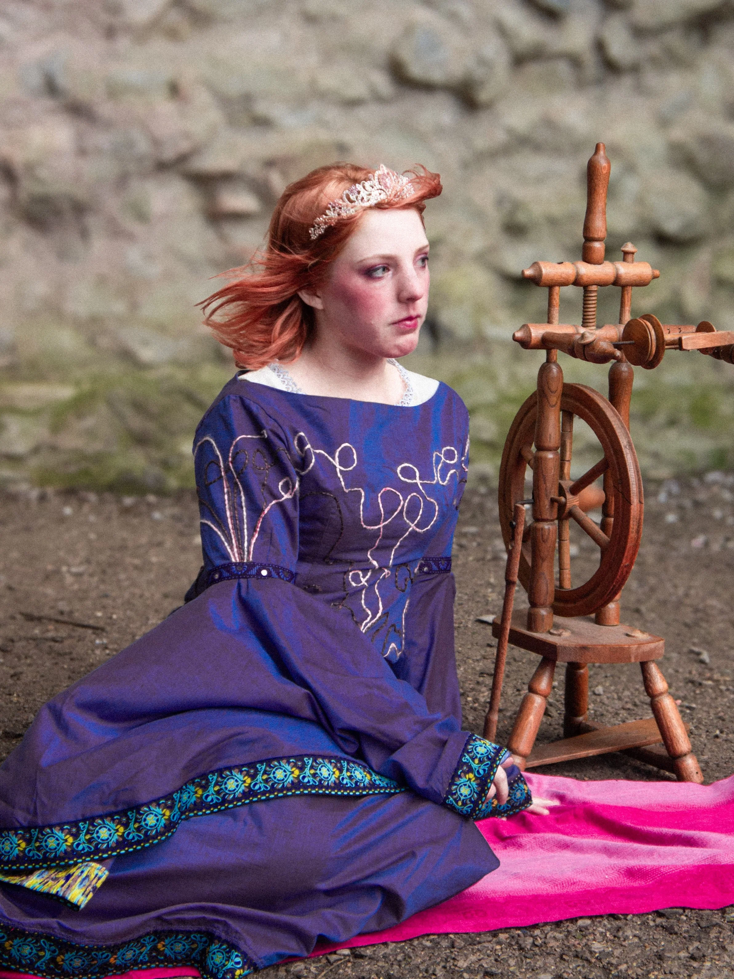 A young woman with red hair wearing a blue dress with embroidery and a lace collar sits on the ground in front of a spinning wheel, looking contemplative.
