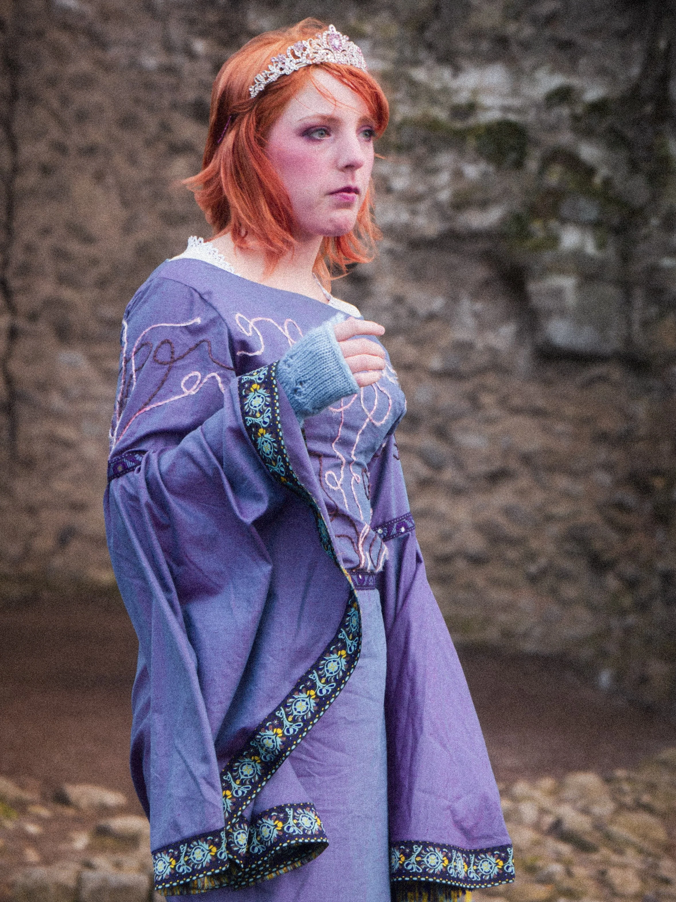 A young woman with red hair wearing a purple dress and a tiara, standing outdoors against a rocky background.
