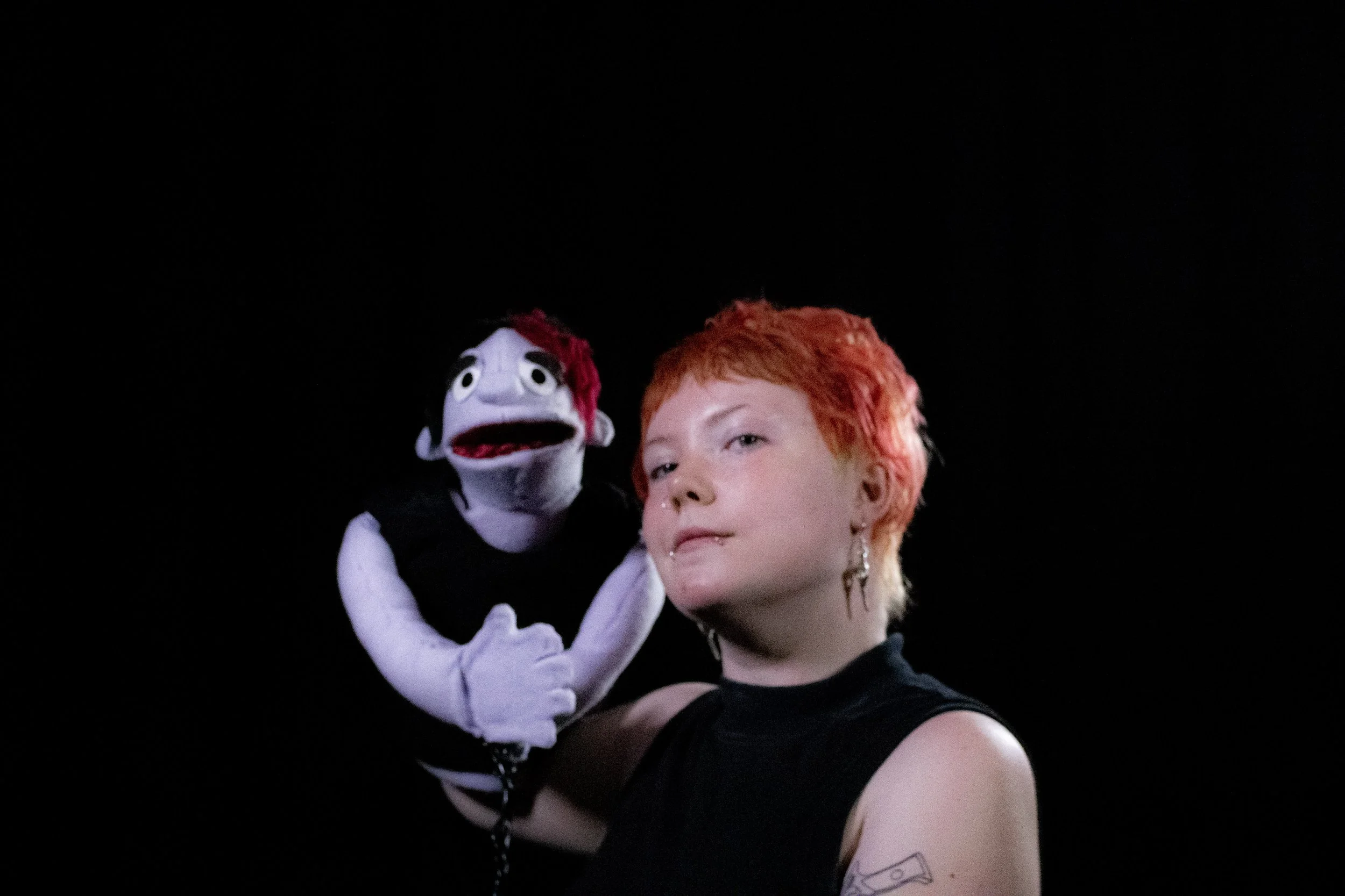 A person with short red hair and earrings holding a puppet with red hair and a painted face against a black background.