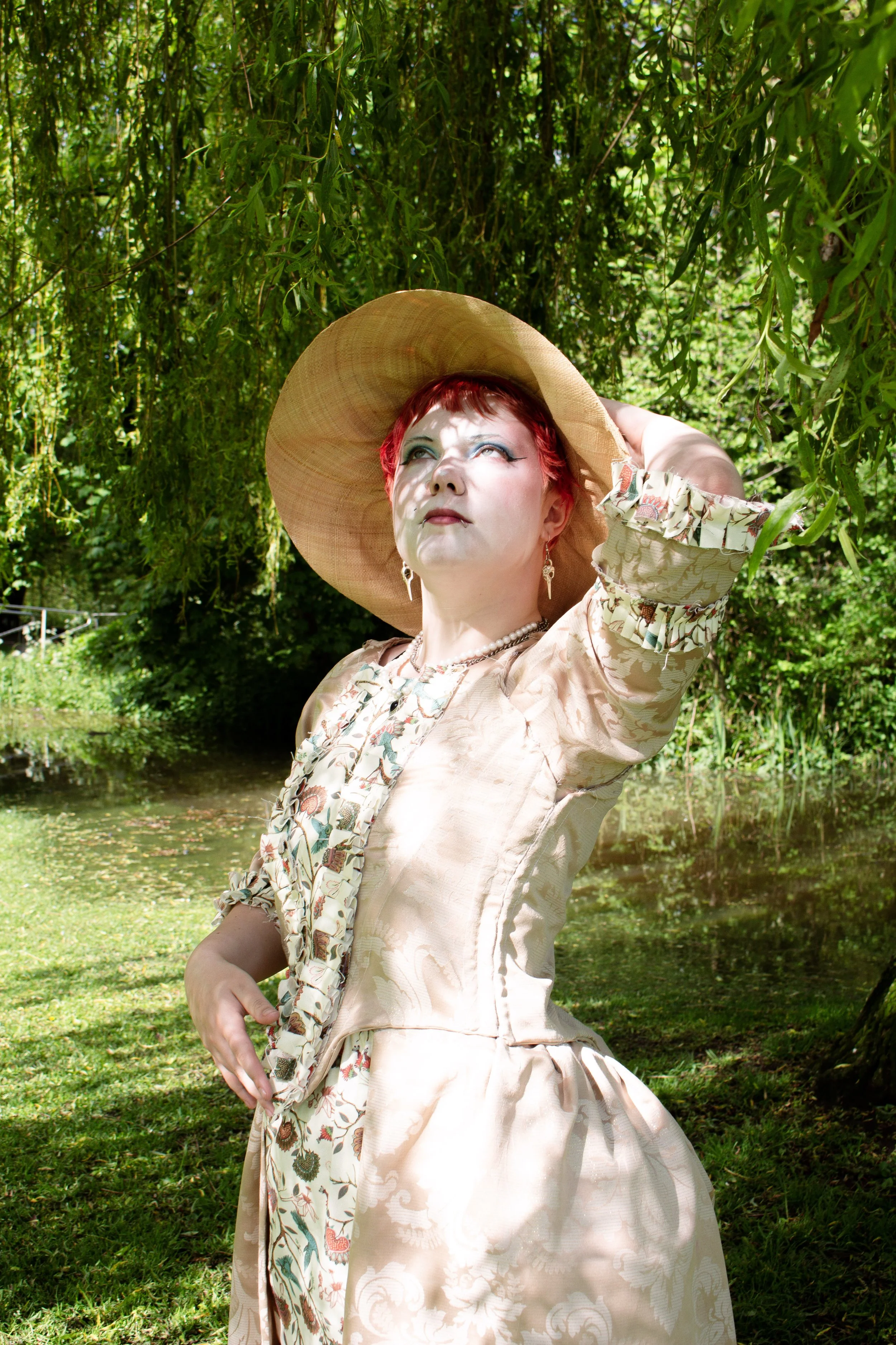 A woman with red hair and pale makeup gazes upward while wearing a large straw hat, standing outdoors near a pond and lush greenery.