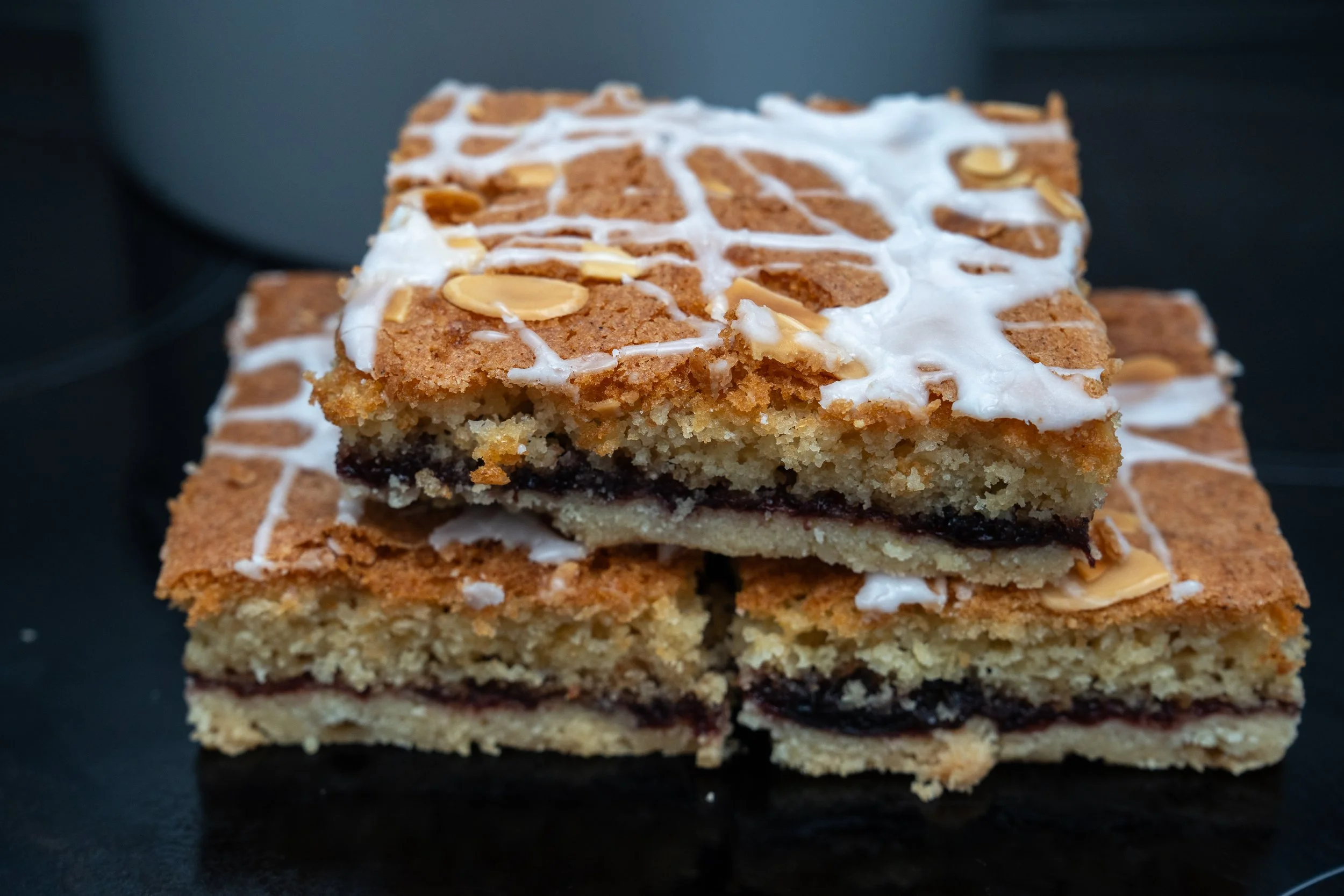 A close-up of a homemade layered dessert bar with a graham cracker crust, dark fruit jam center, and white icing with almond slices on top, resting on a black surface.