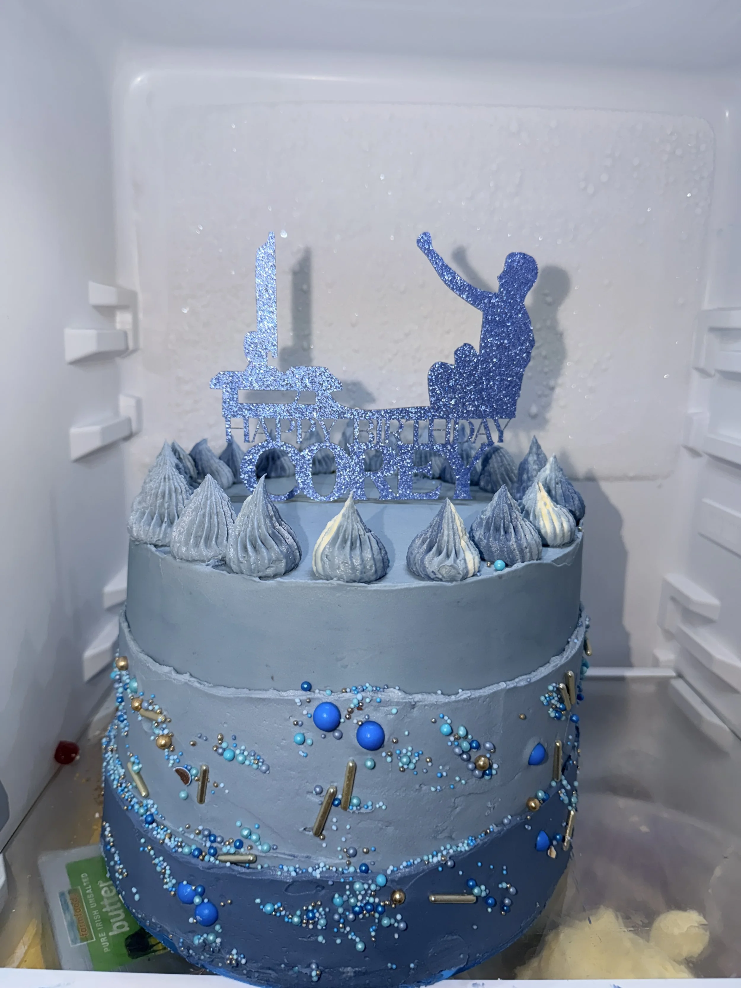 A multi-tiered birthday cake decorated with blue and gold beads and a sparkly blue birthday topper. The topper features a silhouette of a child reaching for a star and the words "Happy Birthday" and "Corey." The cake is inside a refrigerator with a f