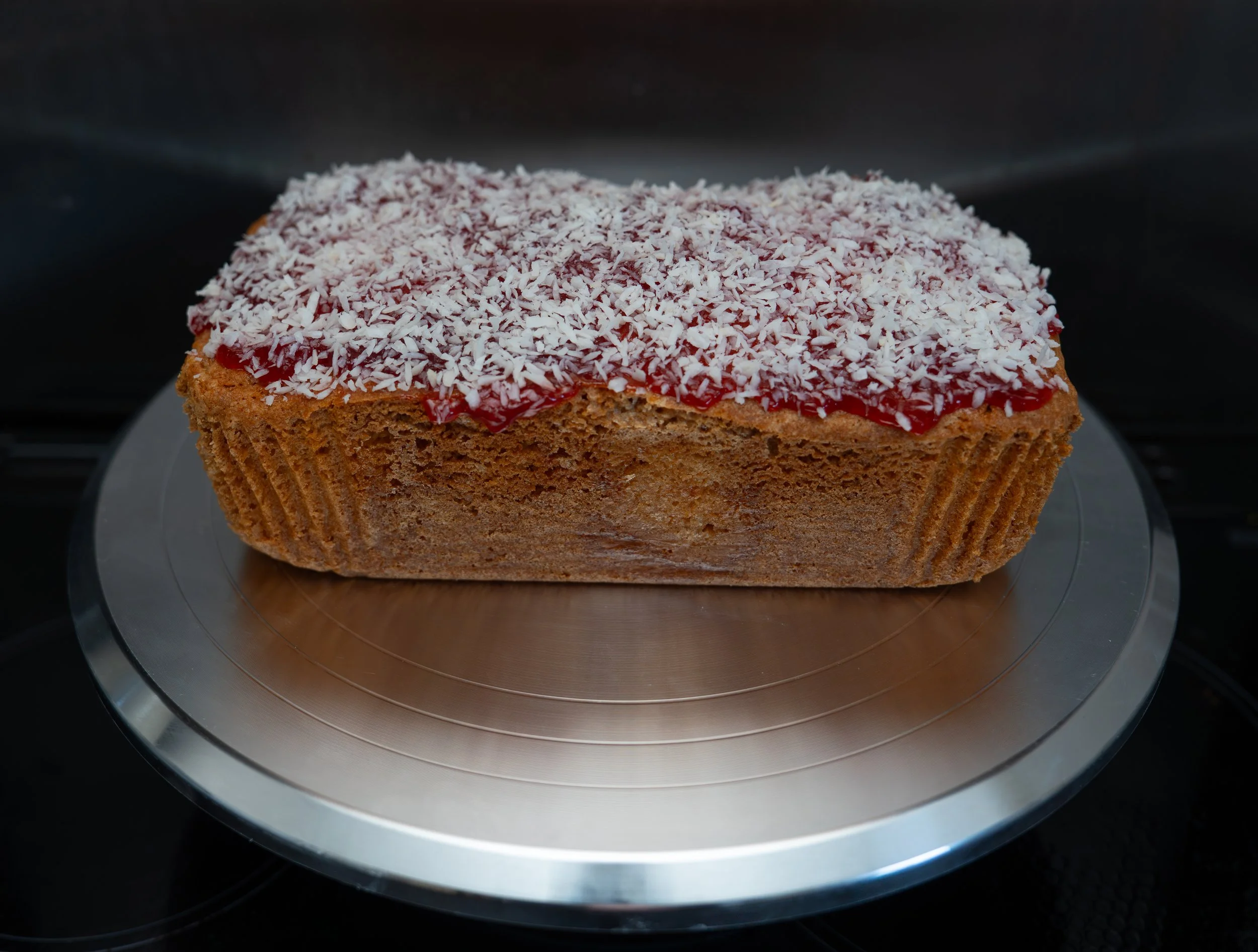 A close-up of a piece of cake with shredded coconut on top, with visible red jam at the edges, placed on a metallic turntable surface.