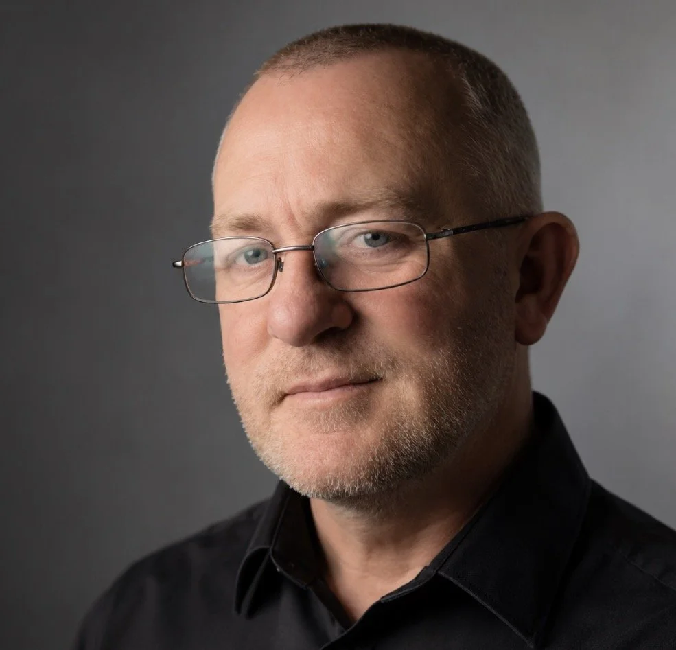 Close-up portrait of a middle-aged man with glasses, short hair, and a slight smile, wearing a black shirt against a gray background.