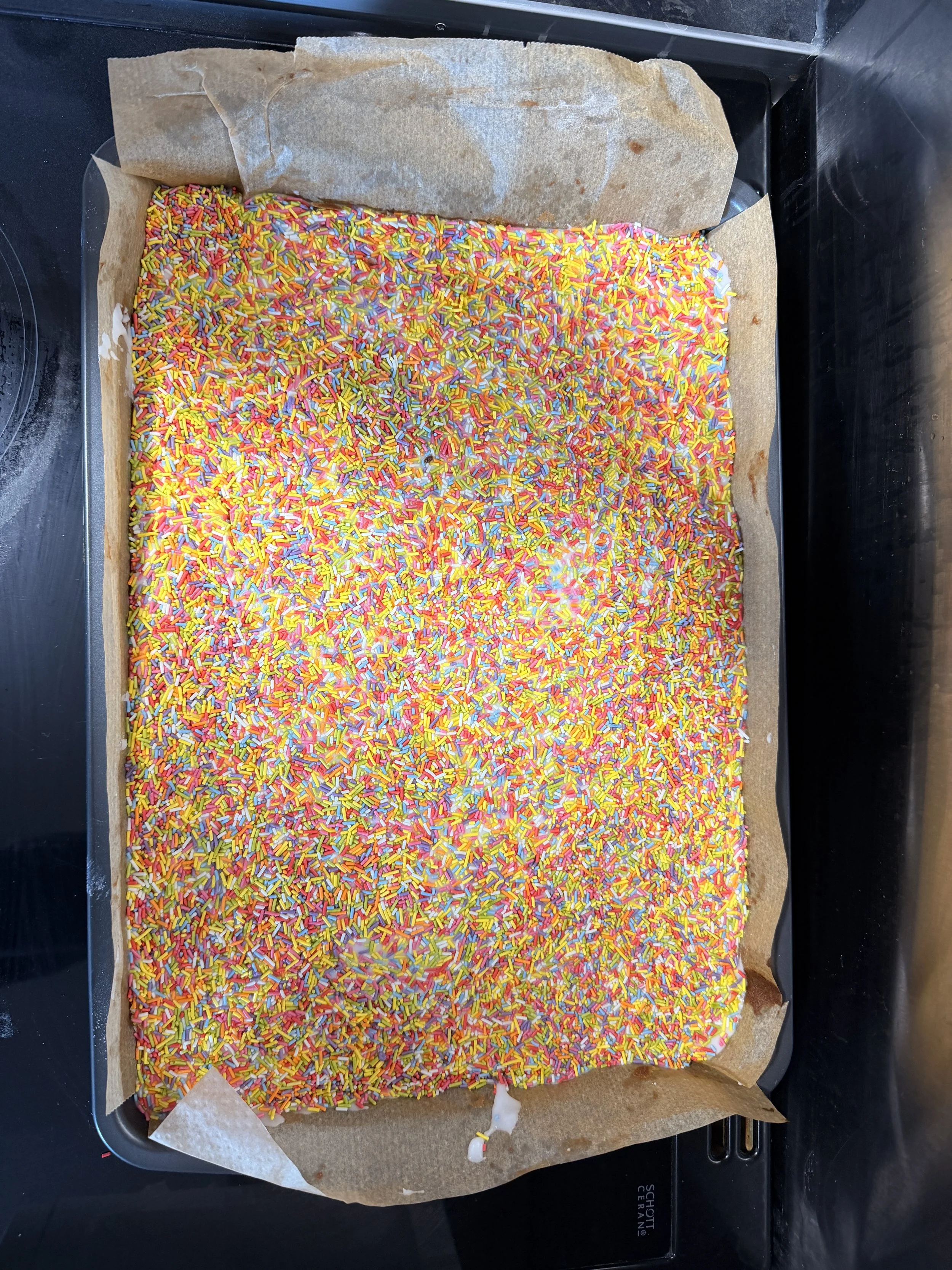 Unbaked rectangular cake with white frosting, covered with multicolored sprinkles, on a baking sheet lined with parchment paper.