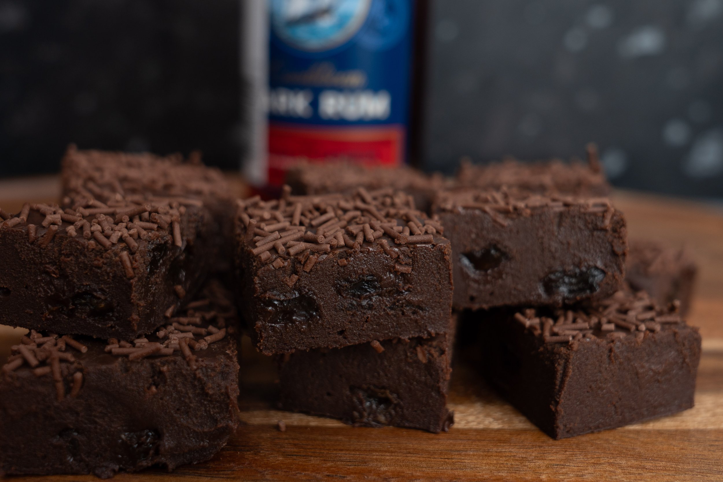 Close-up of chocolate brownies topped with chocolate sprinkles, stacked on a wooden surface with a blurred background.