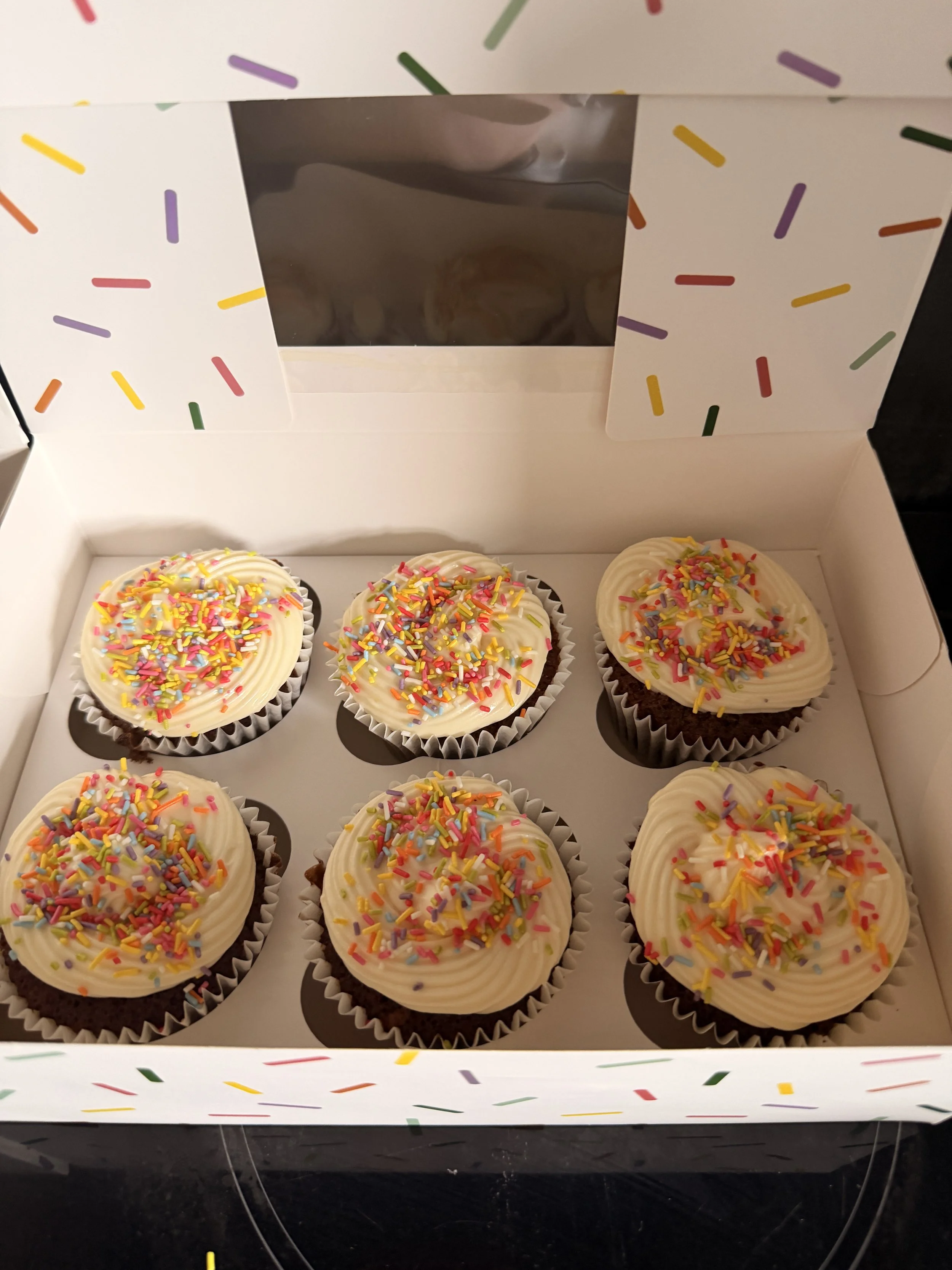 Six chocolate cupcakes with vanilla frosting and colorful sprinkles inside a white box with a clear window and rainbow sprinkles pattern.