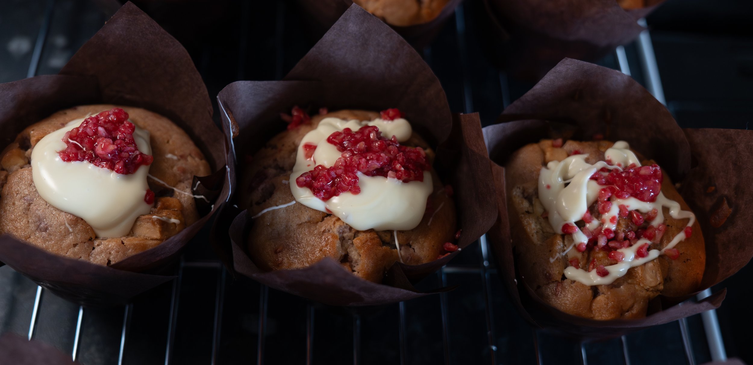 Three muffins with white frosting and red fruit topping on a wire rack