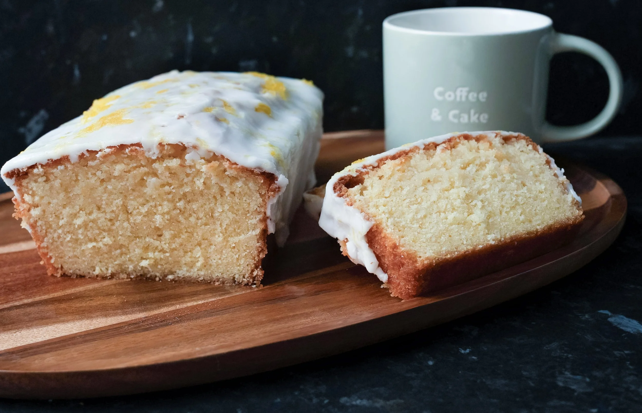 Close-up of slices of lemon drizzle cake on a wooden serving board, with a white mug labeled 'Coffee & Cake' in the background.