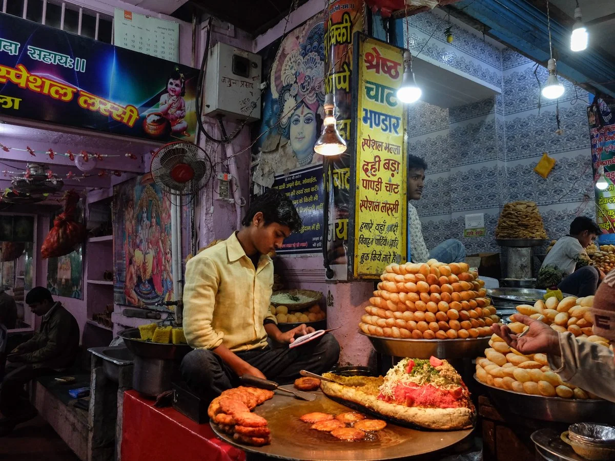 Street food stall in India with fried snacks, eggs, and fried bread, with a young man preparing food and customers nearby. Colorful signs in Hindi and images of deities decorate the stall.
