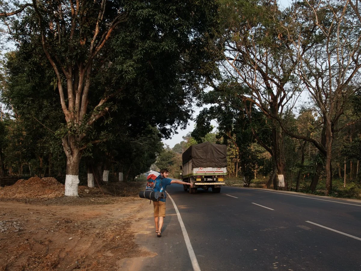 A person with a backpack and rolled-up yoga mat walks along the side of a road lined with trees, as a truck drives by in the opposite direction.