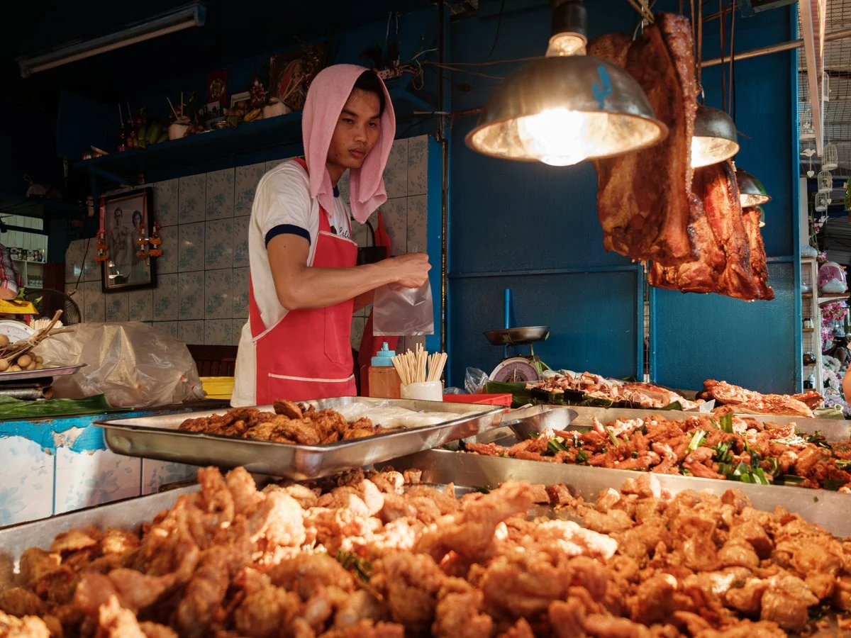 A young man wearing a pink towel on his head and a red apron stands behind a street food stall, preparing food. In front of him are trays of fried chicken and other cooked meats, with large strips of cooked pork hanging above. The stall is decorated with hanging lamps and a blue wall background.