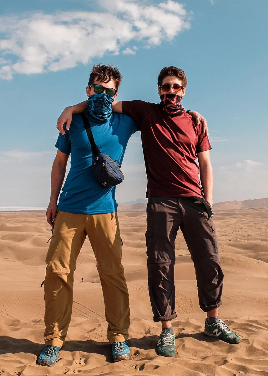 Two young men standing in a desert with sand dunes, wearing casual outdoor clothing, sunglasses, and bandanas covering their faces, posing with their arms around each other.