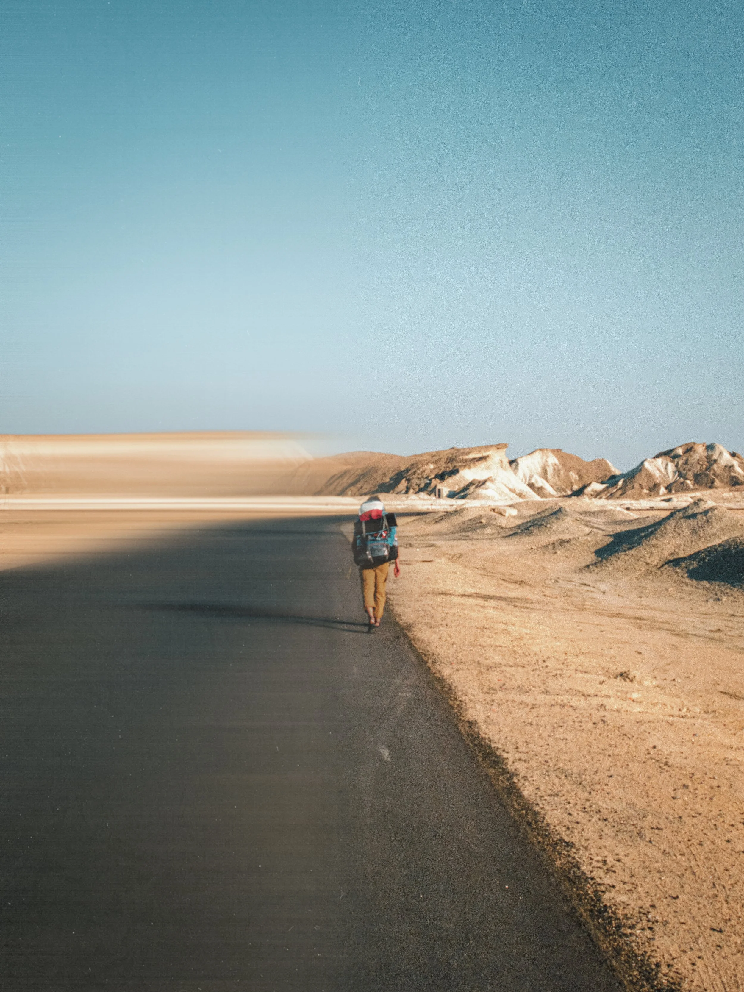 A person walking alone on a paved road in a desert, carrying a backpack, with mountains in the background and a clear blue sky.