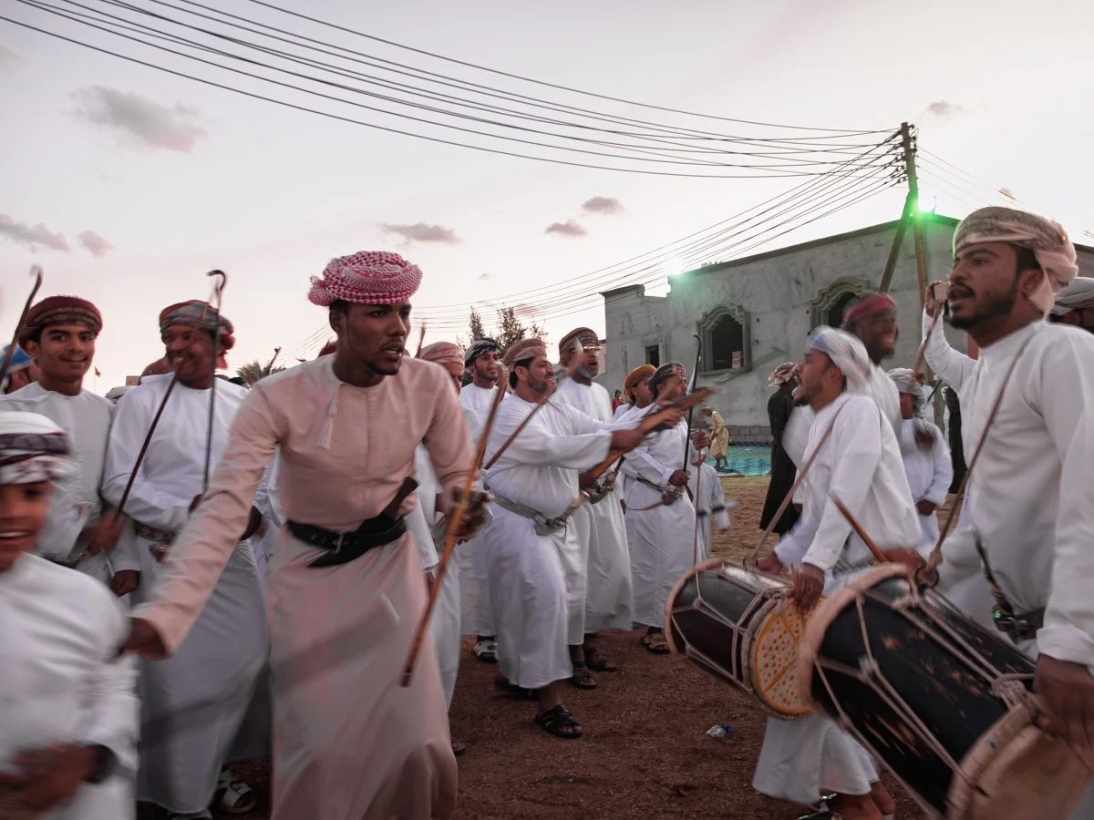 Group of men dressed in traditional Middle Eastern clothing, participating in a folk dance, with some playing drums, outdoors during the evening with a building and power lines in the background.