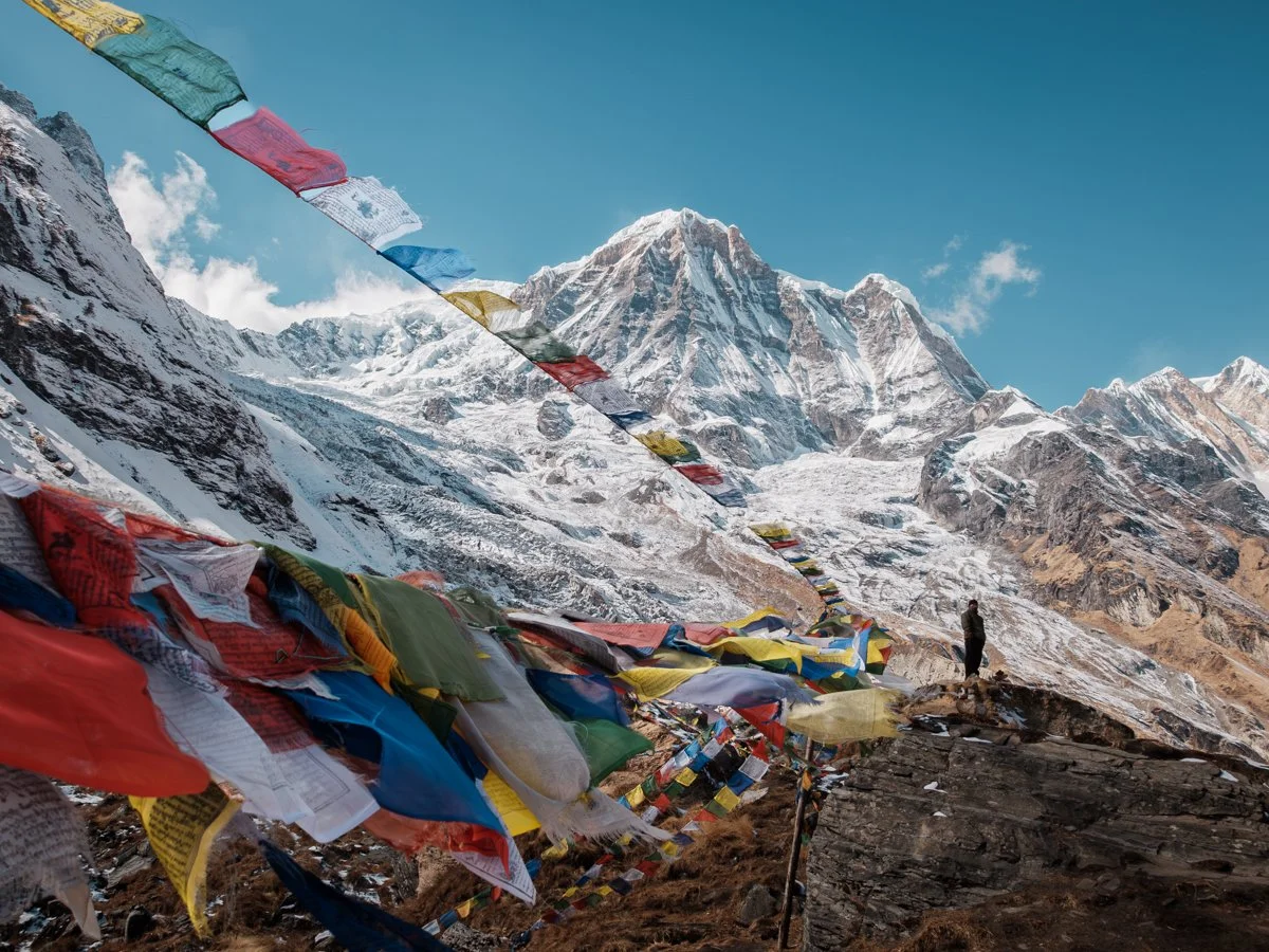 Snow-covered mountains with prayer flags strung across a rocky slope and a person standing, facing the mountains.