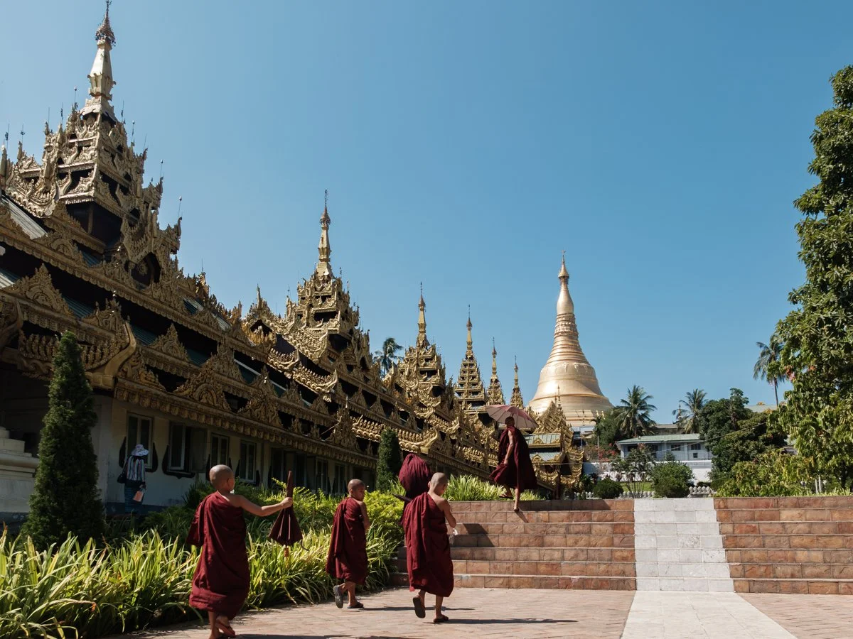 Monks walking near an ornate temple with golden spires and a large stupa in the background, surrounded by trees and greenery.
