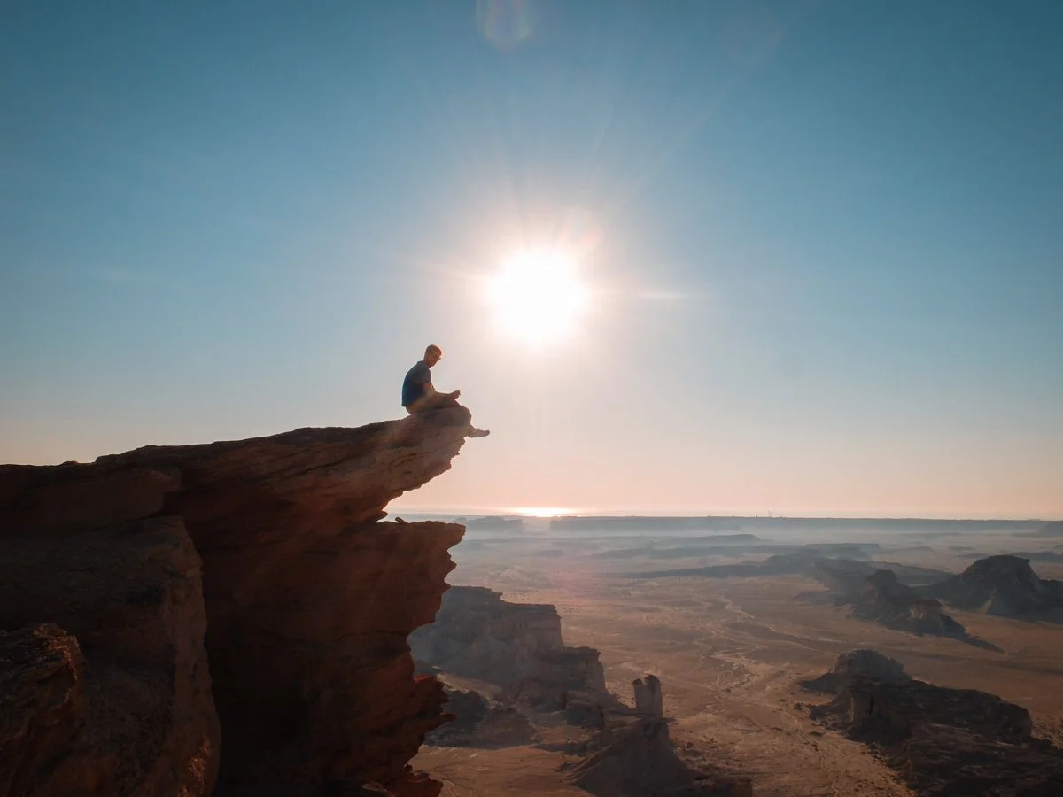 A person sitting on the edge of a large rock formation overlooking a desert landscape during sunset