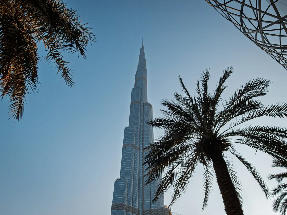 The image shows the Burj Khalifa skyscraper rising into a clear blue sky, framed by palm trees in the foreground.
