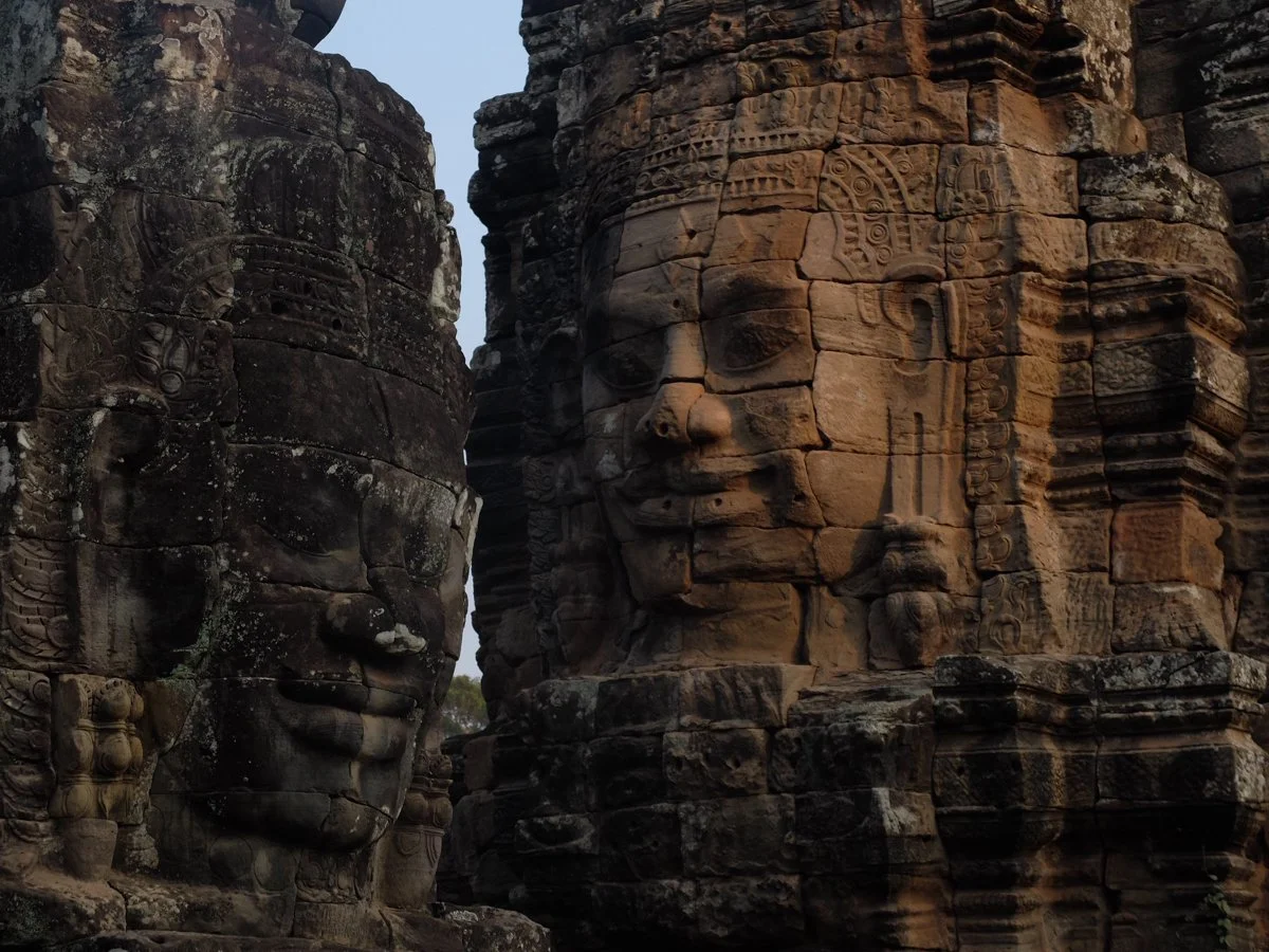 Stone carvings of large human faces on ancient temple towers, likely part of Angkor Wat or a similar Khmer temple complex.