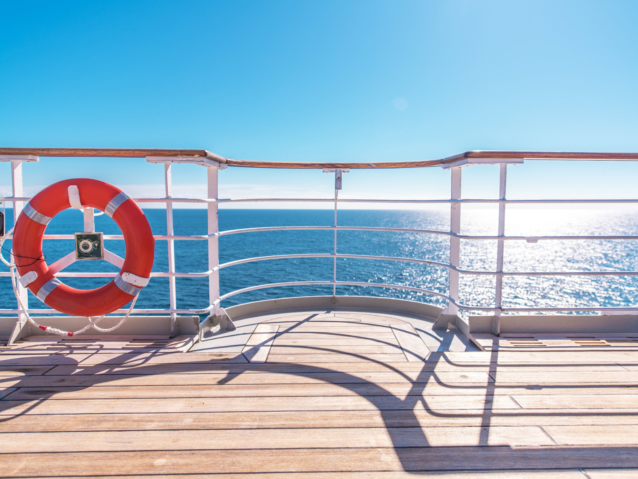 A panoramic view from the wooden deck of a luxury cruise ship, looking out over the vast, calm blue ocean toward a soft horizon at sunset.