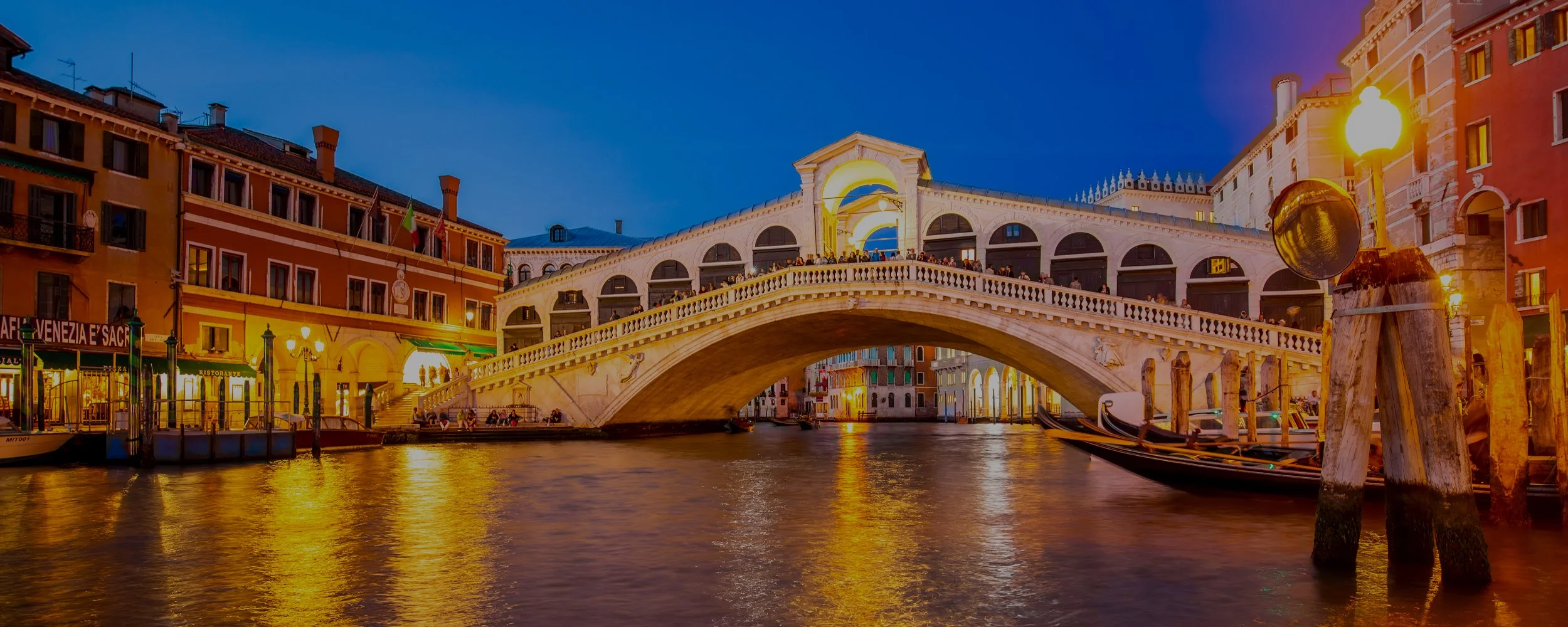 The historic Rialto Bridge in Venice, Italy, illuminated at night with golden lights reflecting on the calm waters of the Grand Canal.
