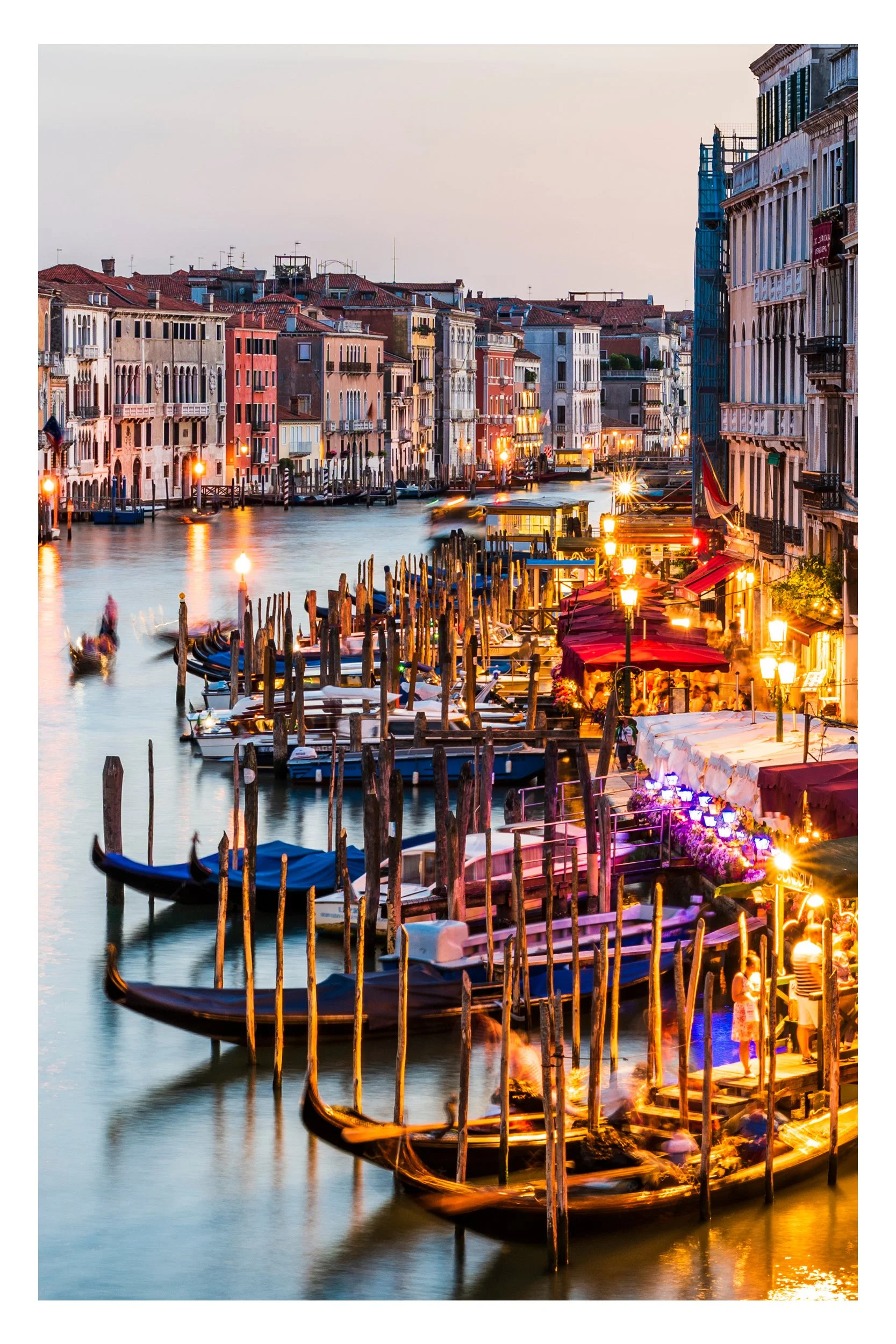 A romantic sunset over the Grand Canal in Venice, Italy, with traditional wooden gondolas moored in the foreground and historic Venetian architecture glowing in the golden evening light.