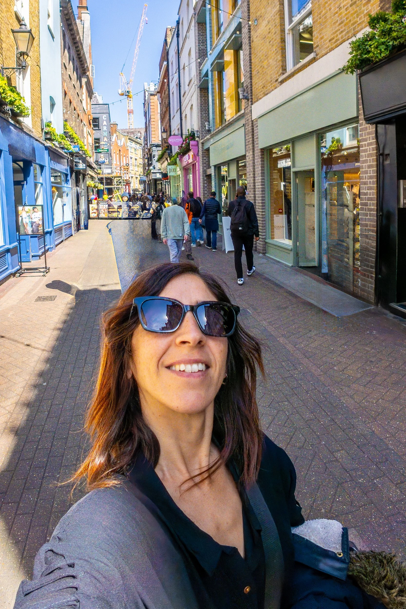 A woman smiling and taking a selfie while walking through a vibrant, busy shopping street in London, England.
