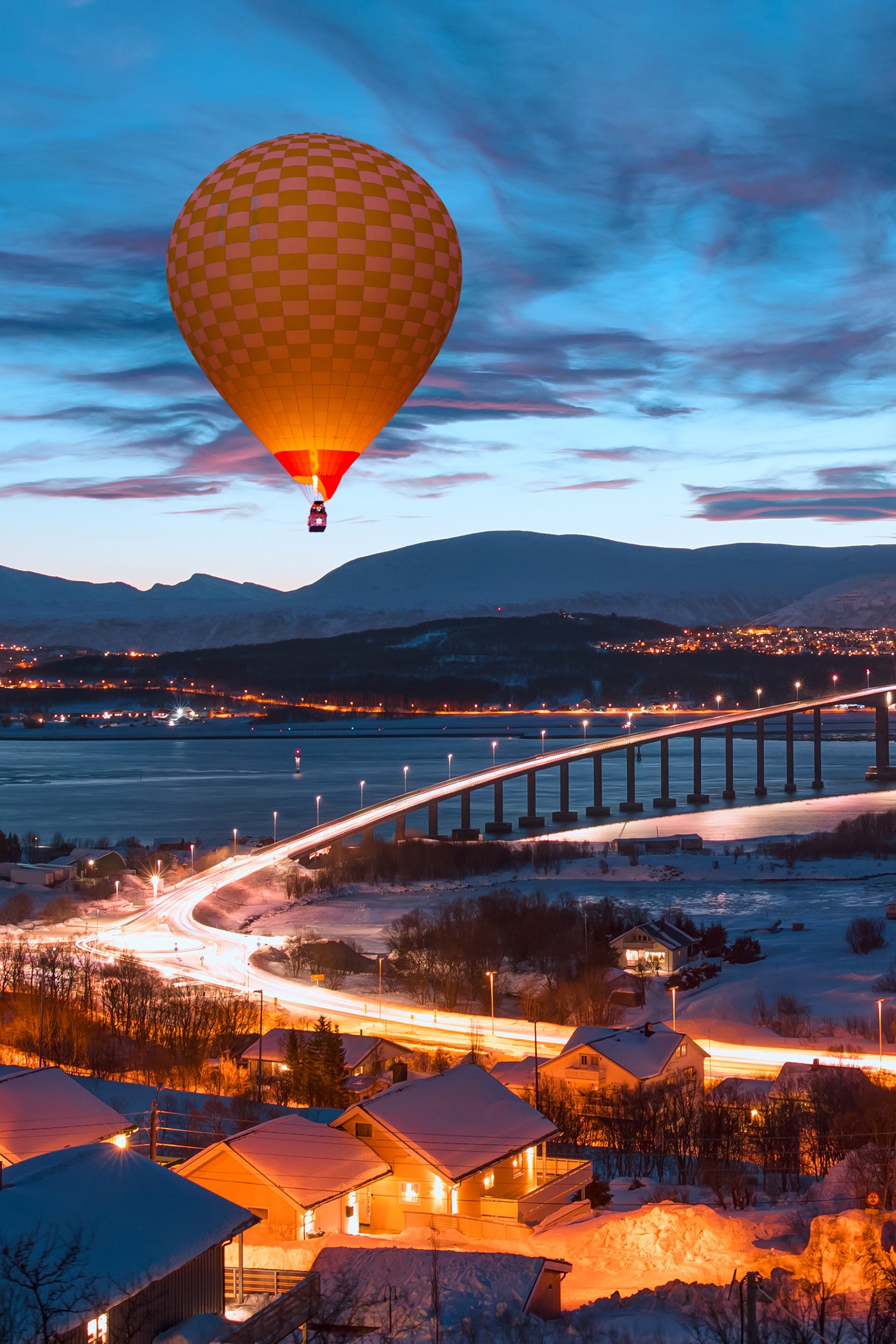 A vibrant hot air balloon drifting over a scenic Norwegian town with traditional architecture and surrounding mountains at sunrise.