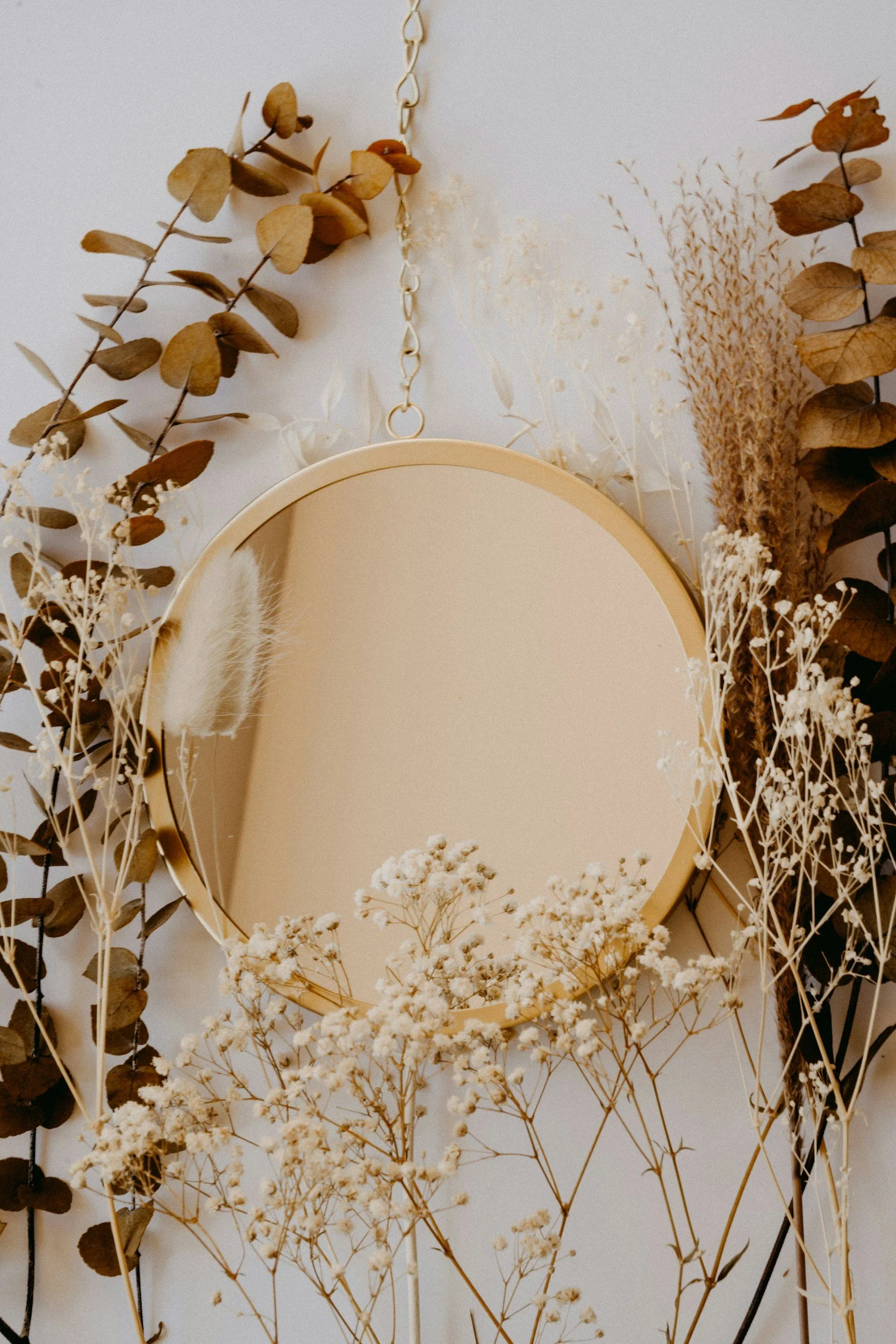 A round mirror hanging from a chain, surrounded by dried brown leaves and white and beige dried flowers against a light wall.