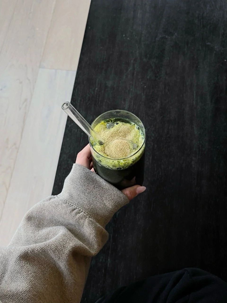 Person holding a glass of green smoothie with a glass straw on dark wooden table.