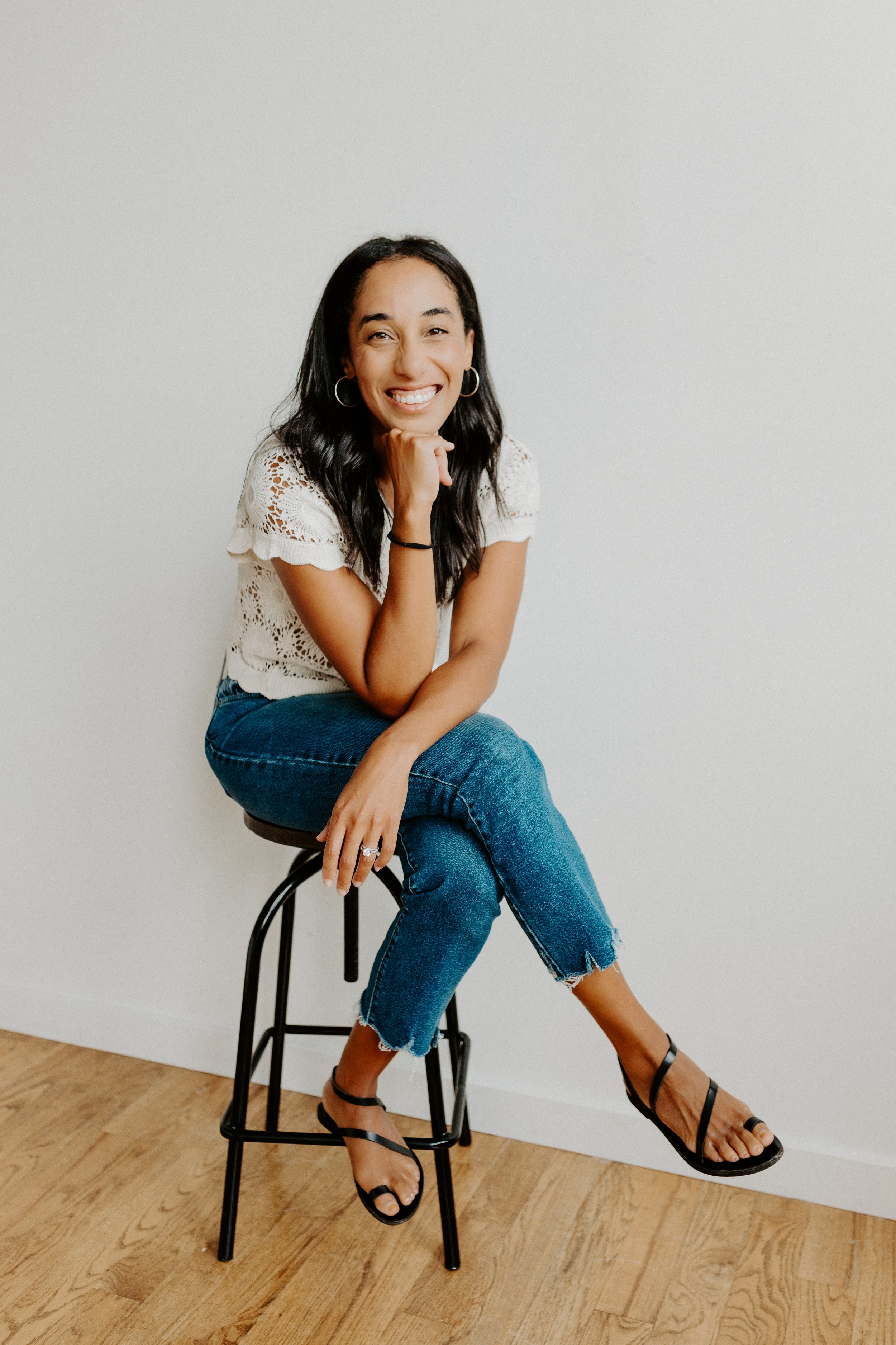 A woman sitting on a black stool with one leg crossed over the other, smiling while wearing a white lace top, blue jeans, and black strappy high-heeled sandals, against a plain white wall and wooden floor
