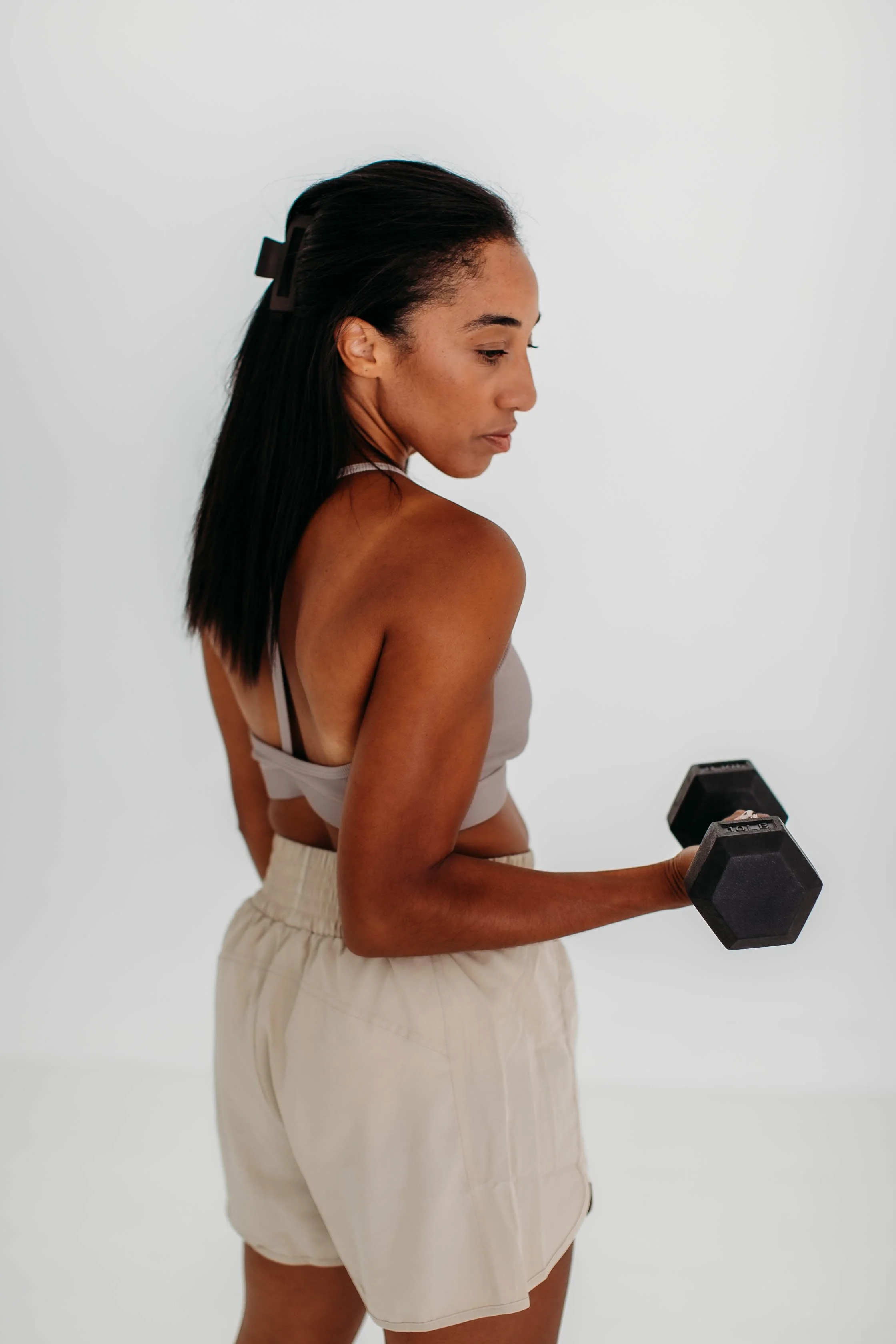 A woman with dark hair in a ponytail, wearing a white tank top and beige shorts, is holding a black dumbbell in her right hand while looking down at her arm against a plain white background.