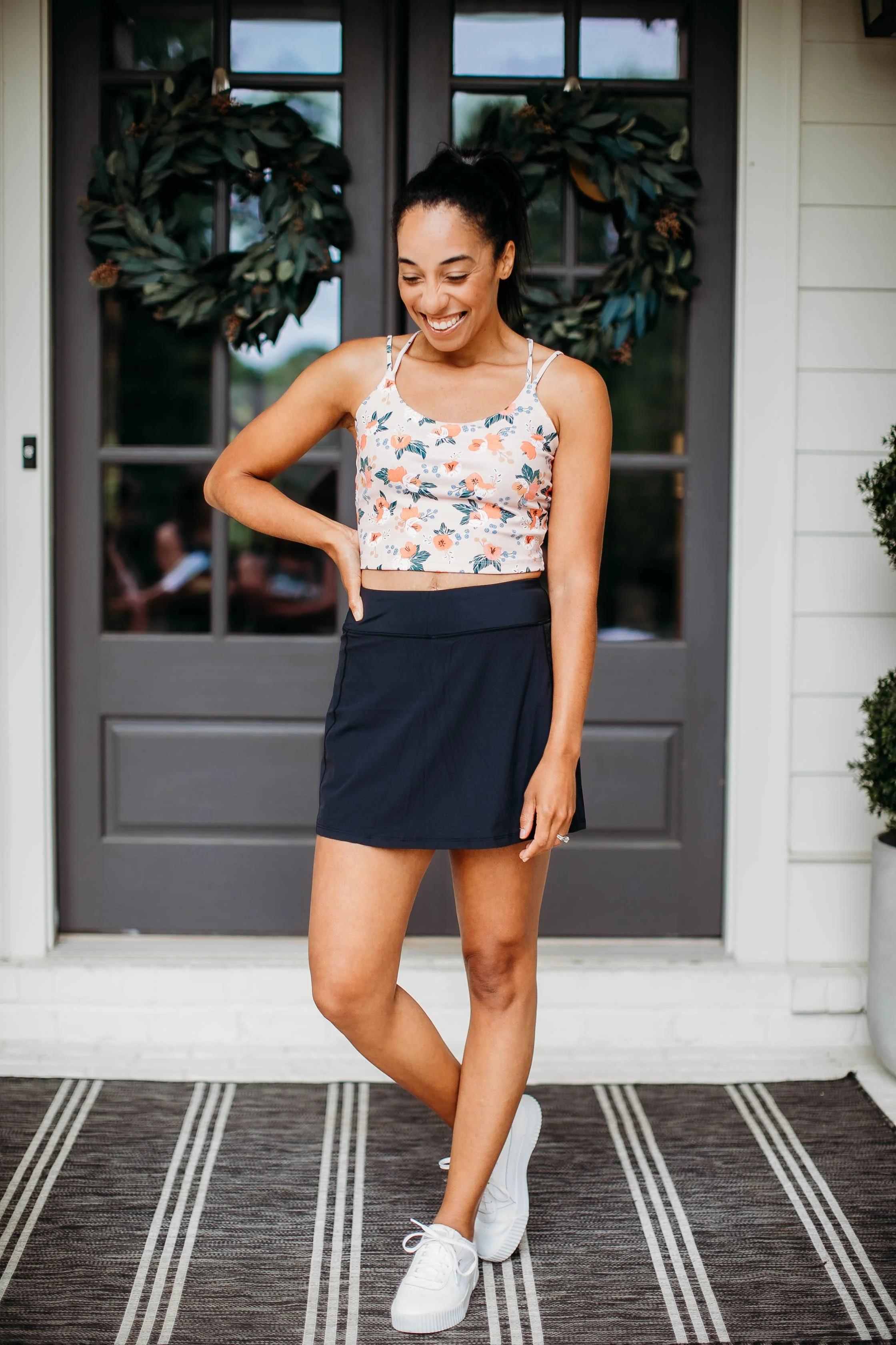 A woman with dark hair in a high ponytail, smiling and looking down, standing on a porch in front of a black door with a leafy wreath, wearing a floral crop top, black skirt, and white sneakers.