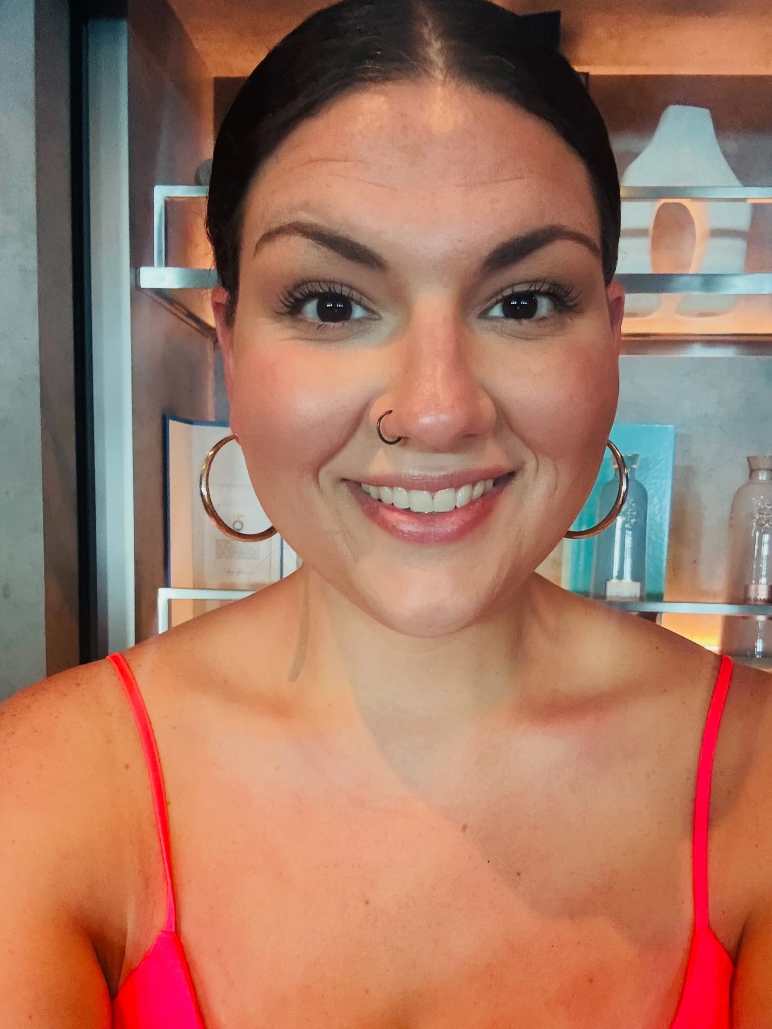 A smiling woman with dark hair, wearing large hoop earrings, a nose ring, and a pink spaghetti strap top, standing in front of a shelf with decorative items.