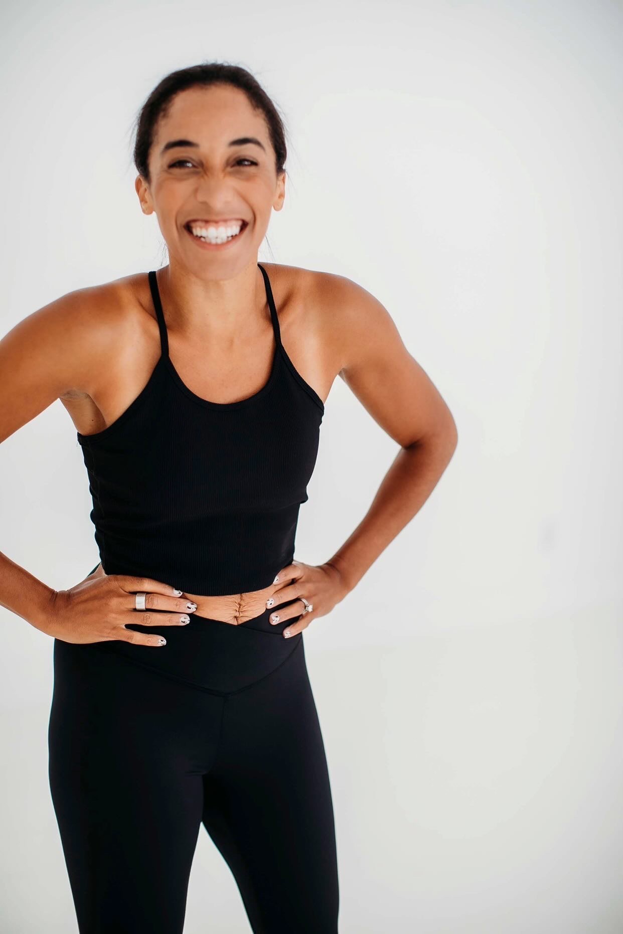 A woman with dark hair in a ponytail, smiling widely, standing with hands on hips against a plain white background. She is wearing a black sports bra and black leggings.