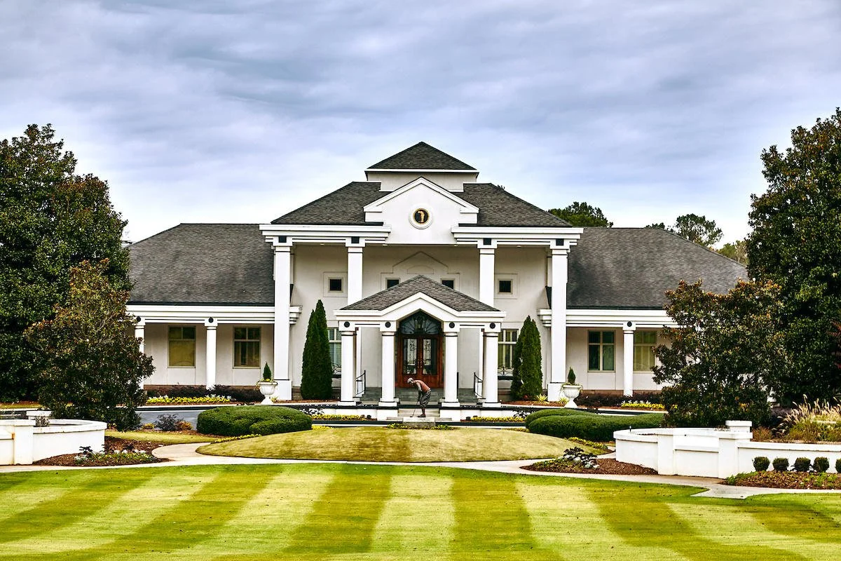 Large white mansion with columns, landscaped yard, and cloudy sky