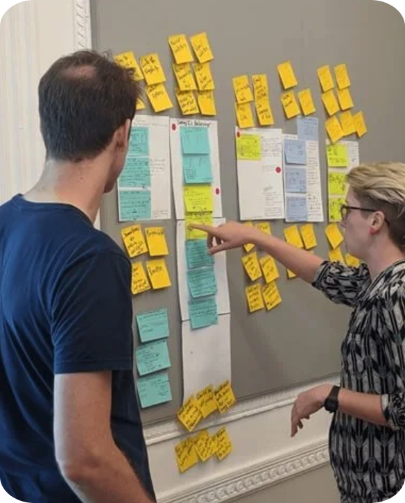 Two people standing near a wall with numerous yellow and blue sticky notes and papers organized in columns, engaged in a discussion or planning session.