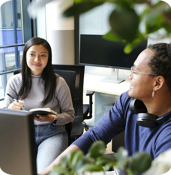 A woman sitting in an office, holding a notebook and pen, smiling while talking to a man wearing headphones, in front of a computer monitor and window with city view.