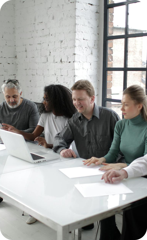 Group of five diverse people sitting at a white table with a laptop and papers, in a modern office with white brick walls and large windows.