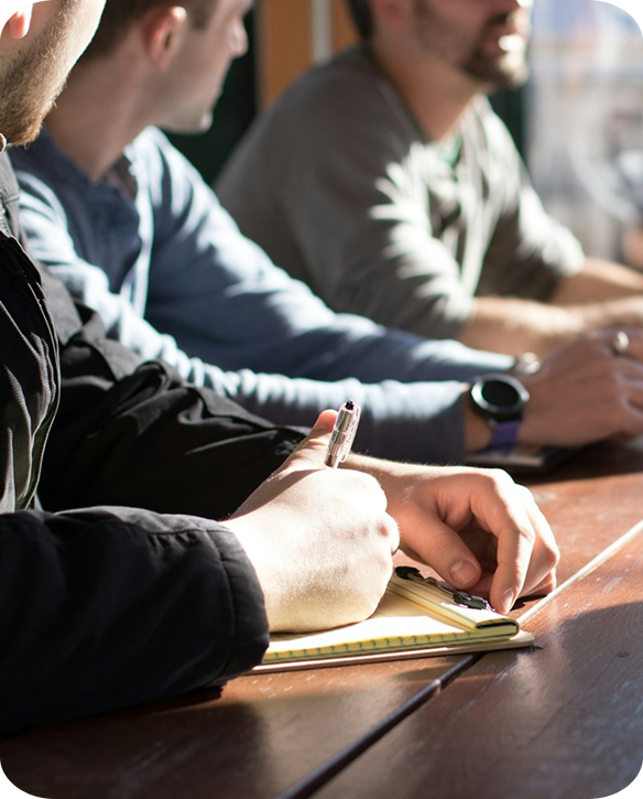 Three people sitting at a wooden table, with one person in the foreground writing on a notepad, and two others sitting before him, possibly in a meeting or discussion setting.