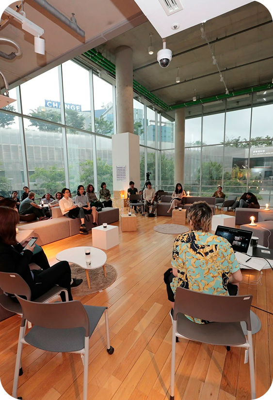 A group of people sitting on sofas and chairs in a modern indoor space with large glass windows, some using laptops and phones, participating in a meeting or event.