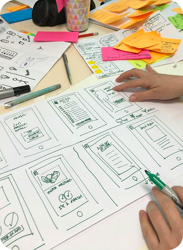 A person is sketching wireframes of a mobile app interface on large paper, with various sketches, notes, and colored sticky notes scattered on the table.