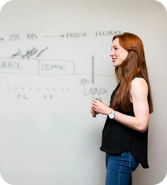 A woman with long red hair and a black sleeveless top standing next to a whiteboard with notes and diagrams.