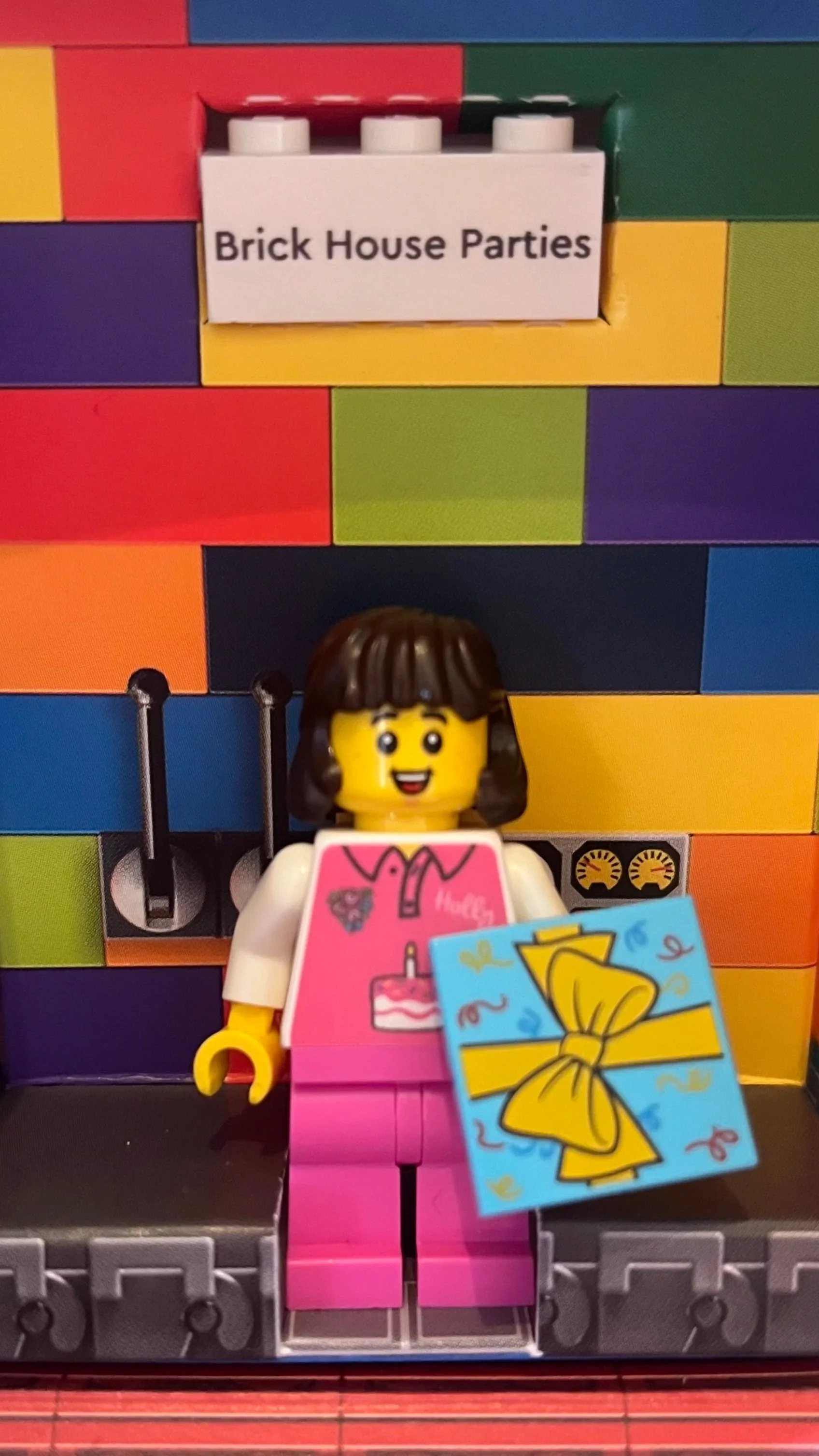 Lego woman holding a gift card standing in front of a colorful Lego background with a sign that reads 'Brick House Parties'.