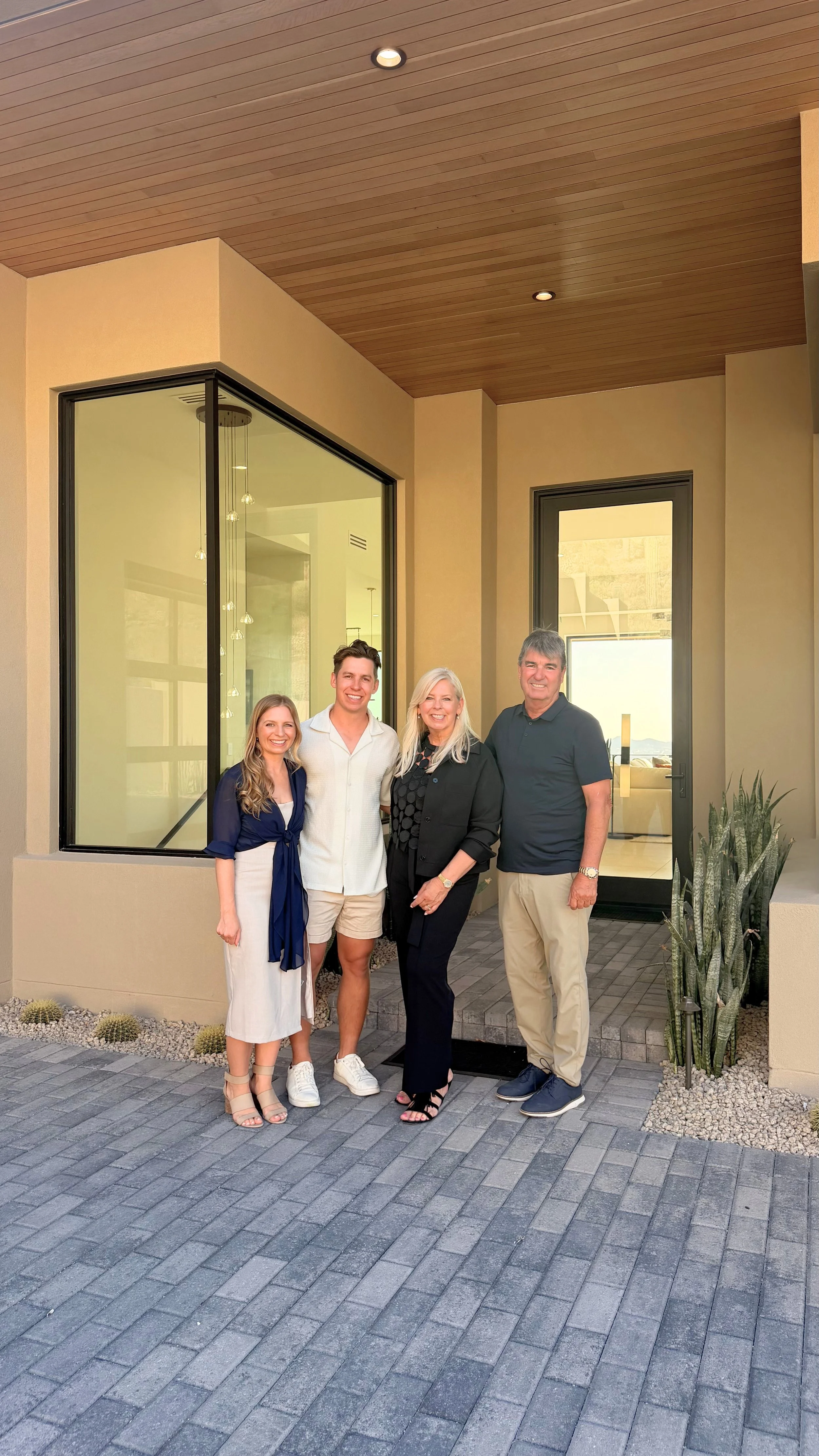 Four people standing outside of a modern house, smiling for the camera, with a large window and a glass door behind them, and a potted plant on the right side.