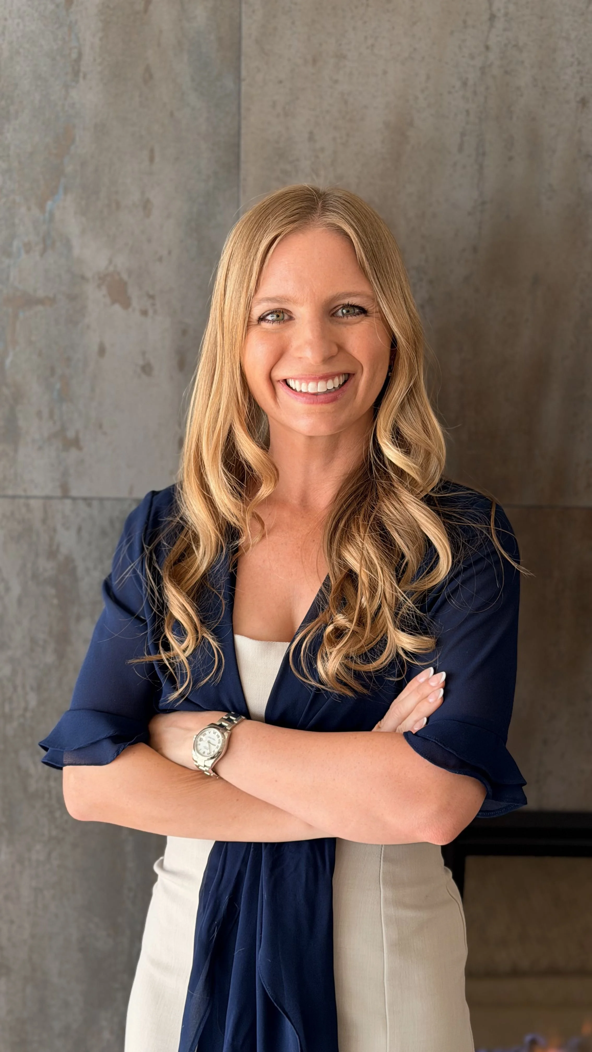 A smiling woman with long wavy blonde hair, crossed arms, wearing a navy blazer, beige skirt, and a wristwatch, standing in front of a plain textured wall.