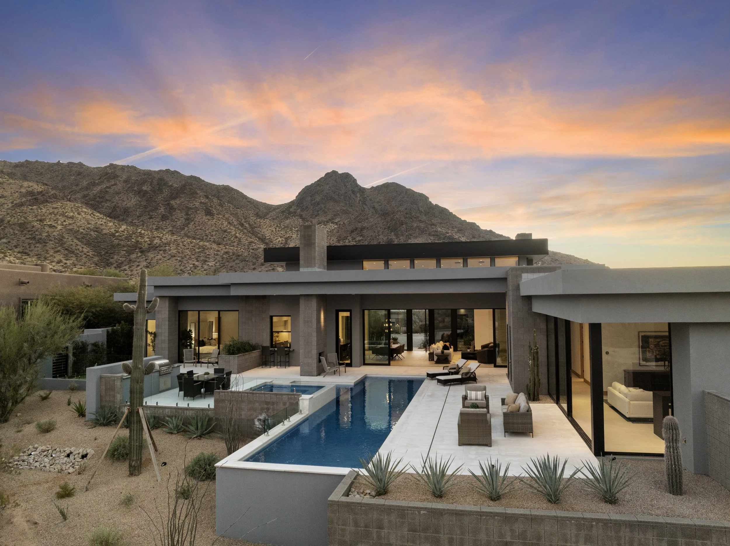 Modern house with large glass windows overlooking a mountainous desert landscape at sunset. The backyard has a swimming pool, outdoor seating, and desert plants.