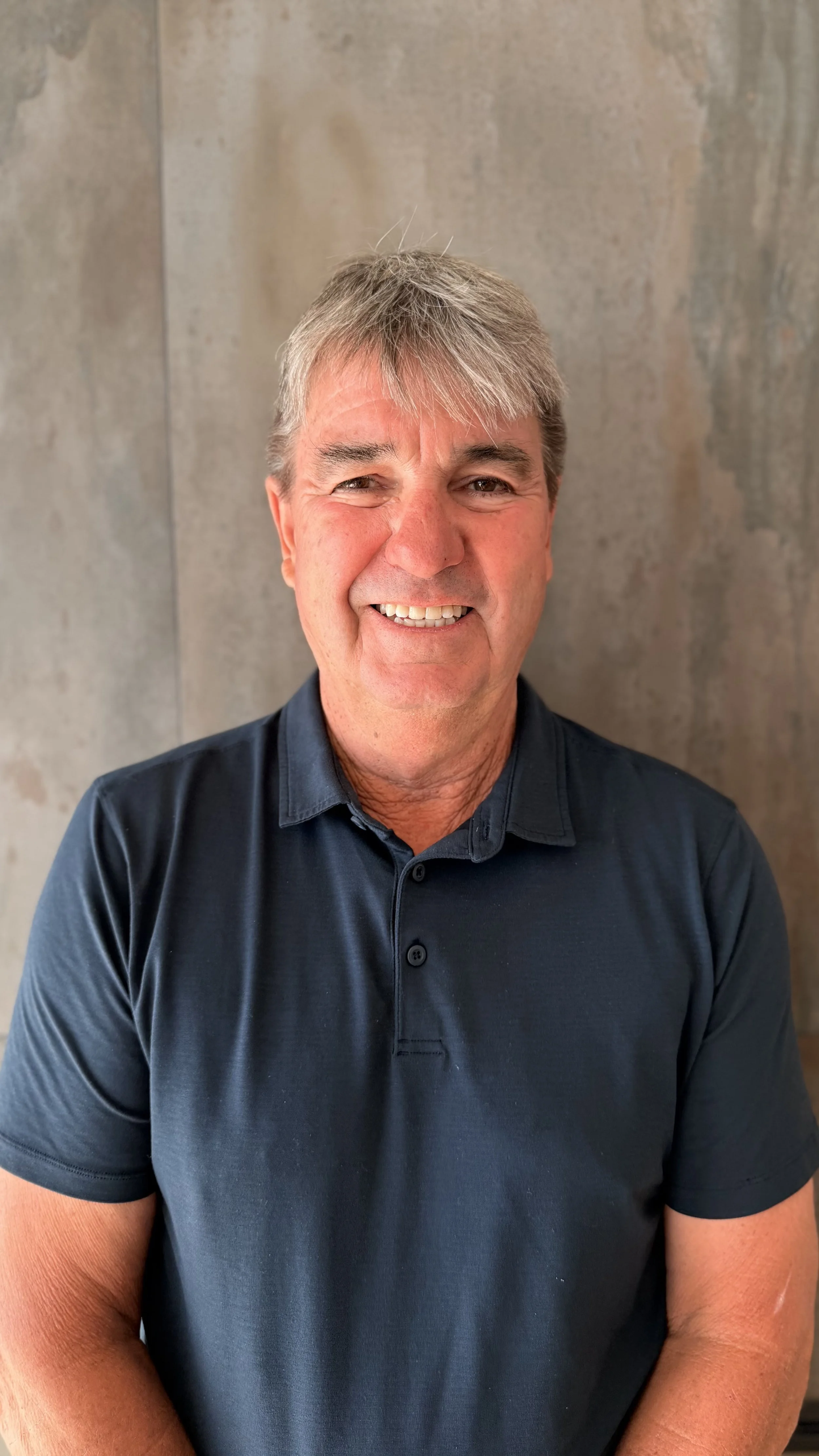 Smiling man with gray hair wearing a dark blue collared shirt against a textured gray background.