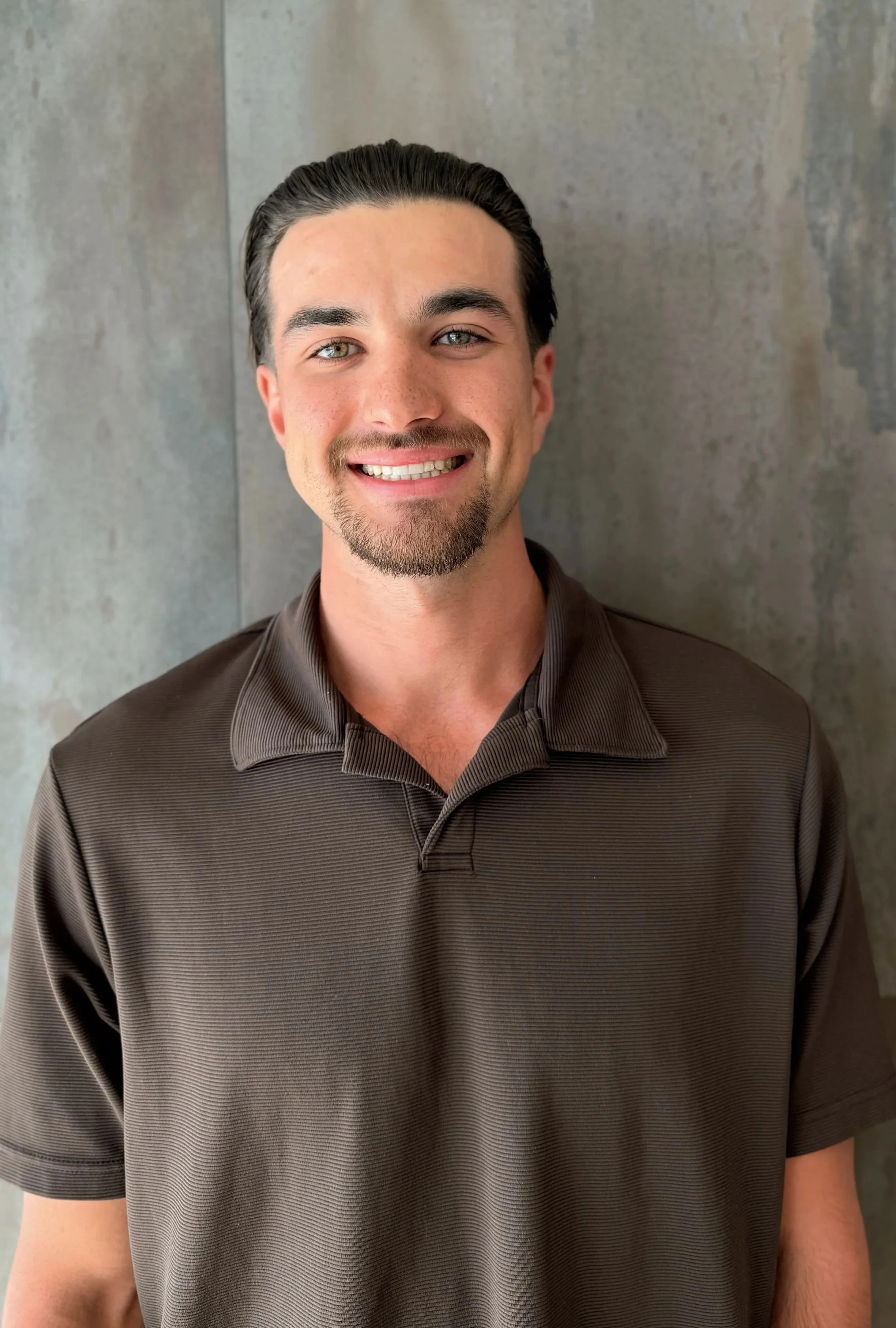 A man with dark hair slicked back, light blue eyes, and a goatee, smiling in front of a gray textured wall. He is wearing a dark brown collared shirt.